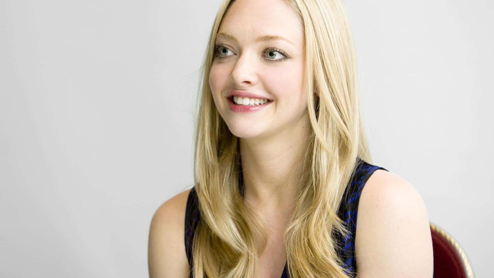 Amanda Seyfried 桌布 壁紙 — 一位微笑的金髮波浪長髮女性，身穿深色圖案上衣，坐在純白背景前。