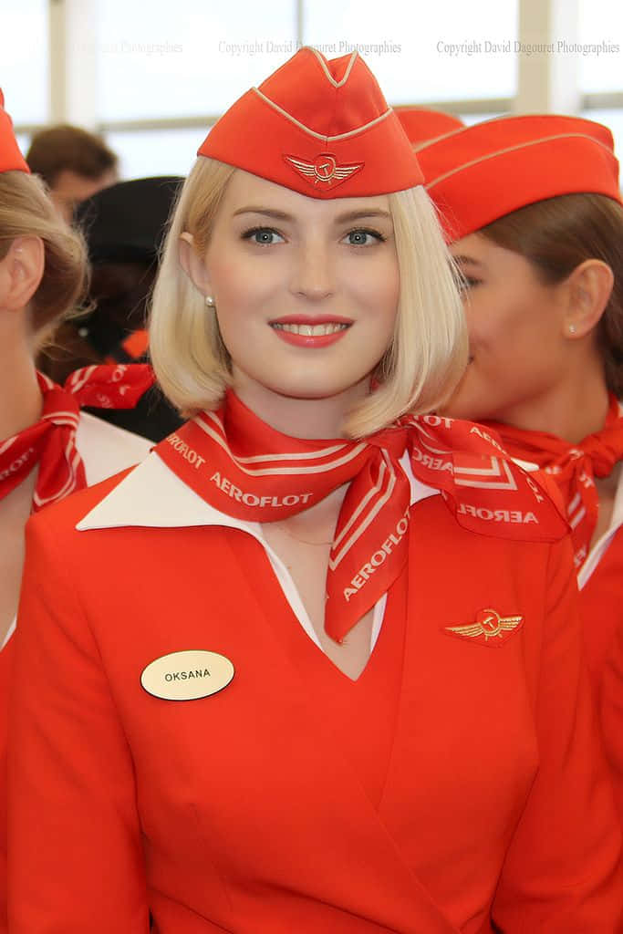 Aircraft 桌布 壁紙 — 一位微笑的金髮空姐穿著鮮紅色Aeroflot制服，搭配相配的帽子和圍巾，站在明亮的室內環境中。