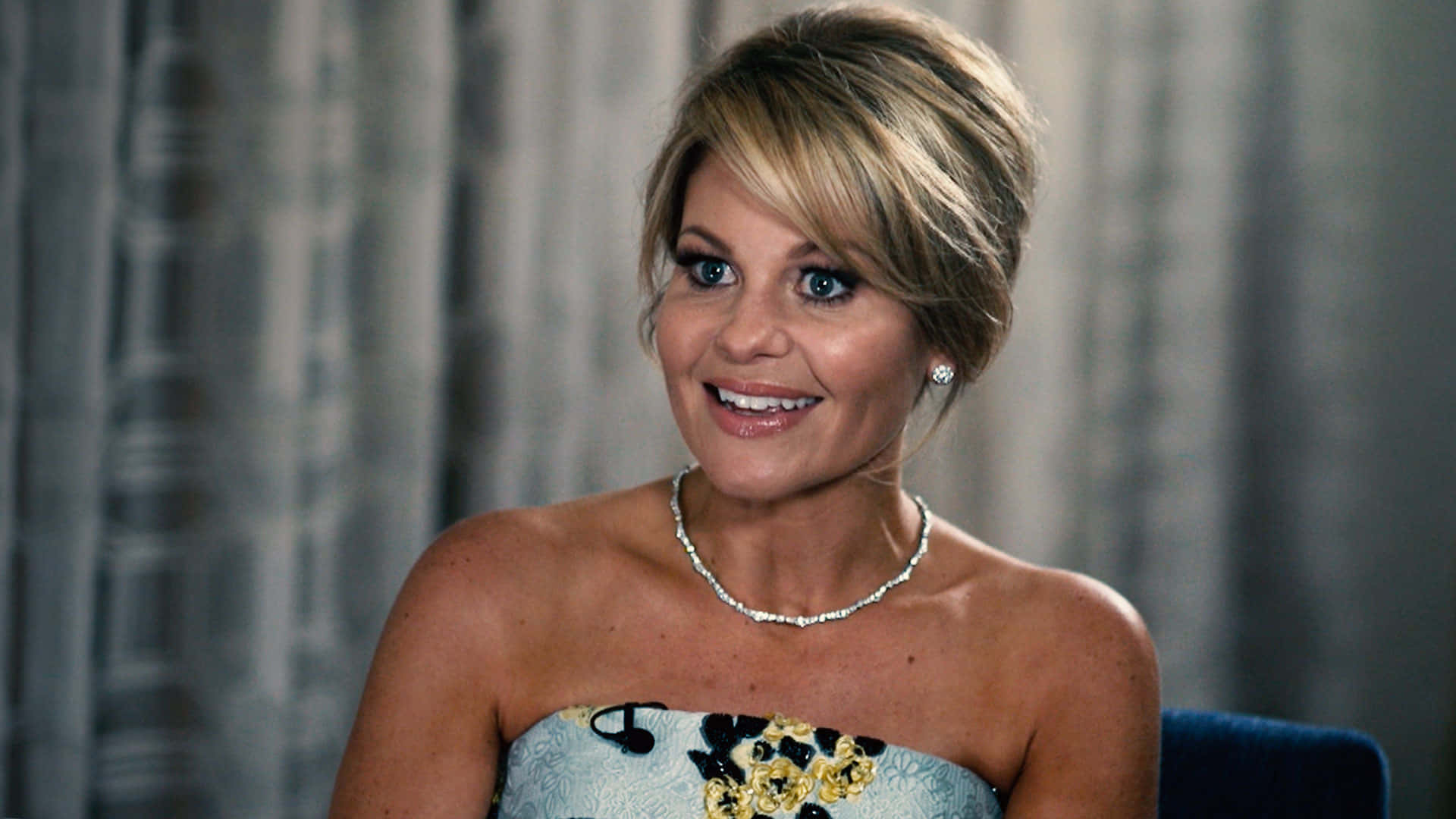 candace cameron bure 桌布 壁紙 — 一位微笑的金髮藍眼女性，穿著無肩帶花卉連衣裙和珍珠首飾，背景是灰色窗簾。