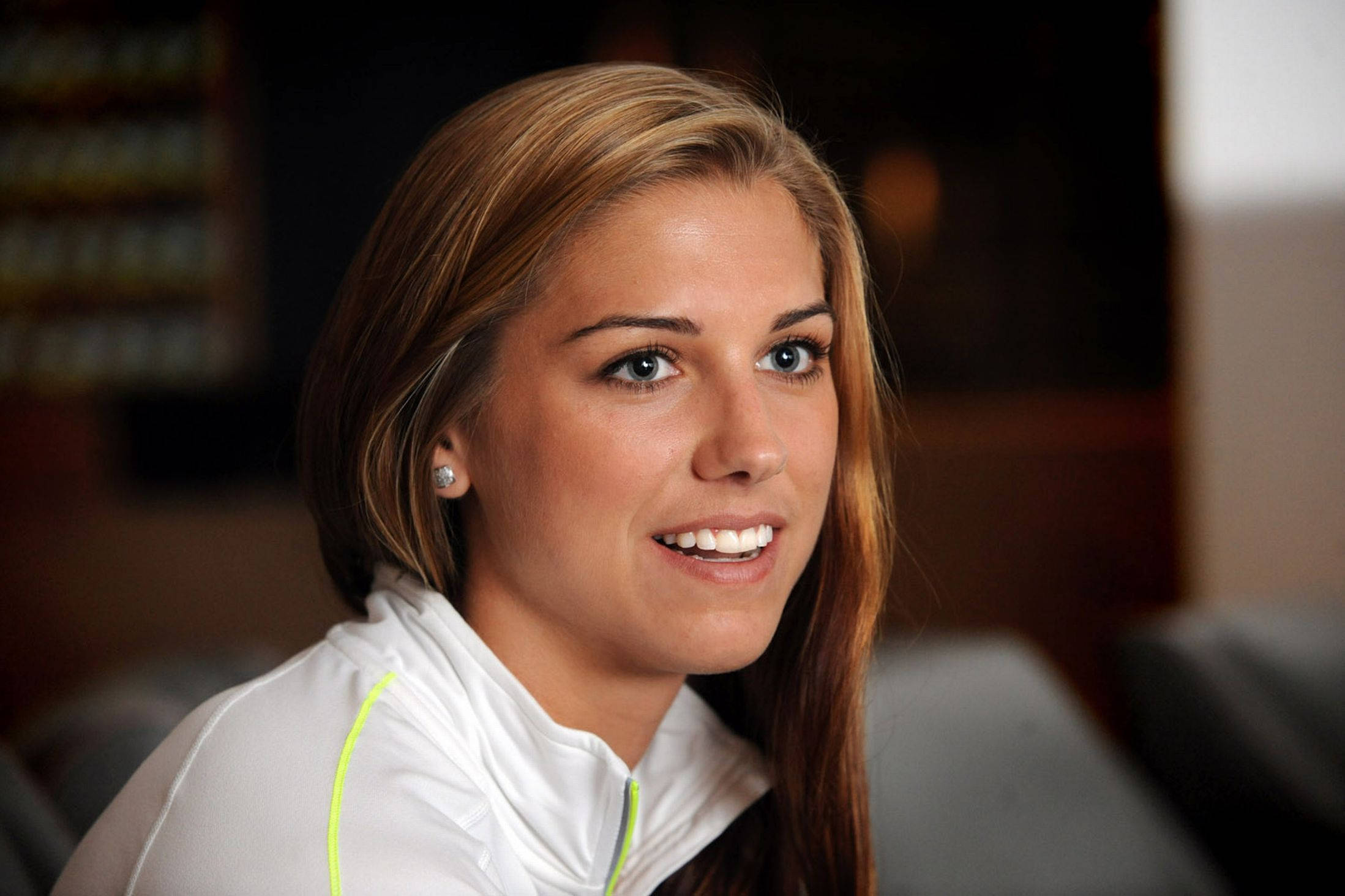 Alex Morgan 桌布 壁紙 — 一位微笑的金髮藍眼女性穿著帶有螢光綠色點綴的白色運動外套。