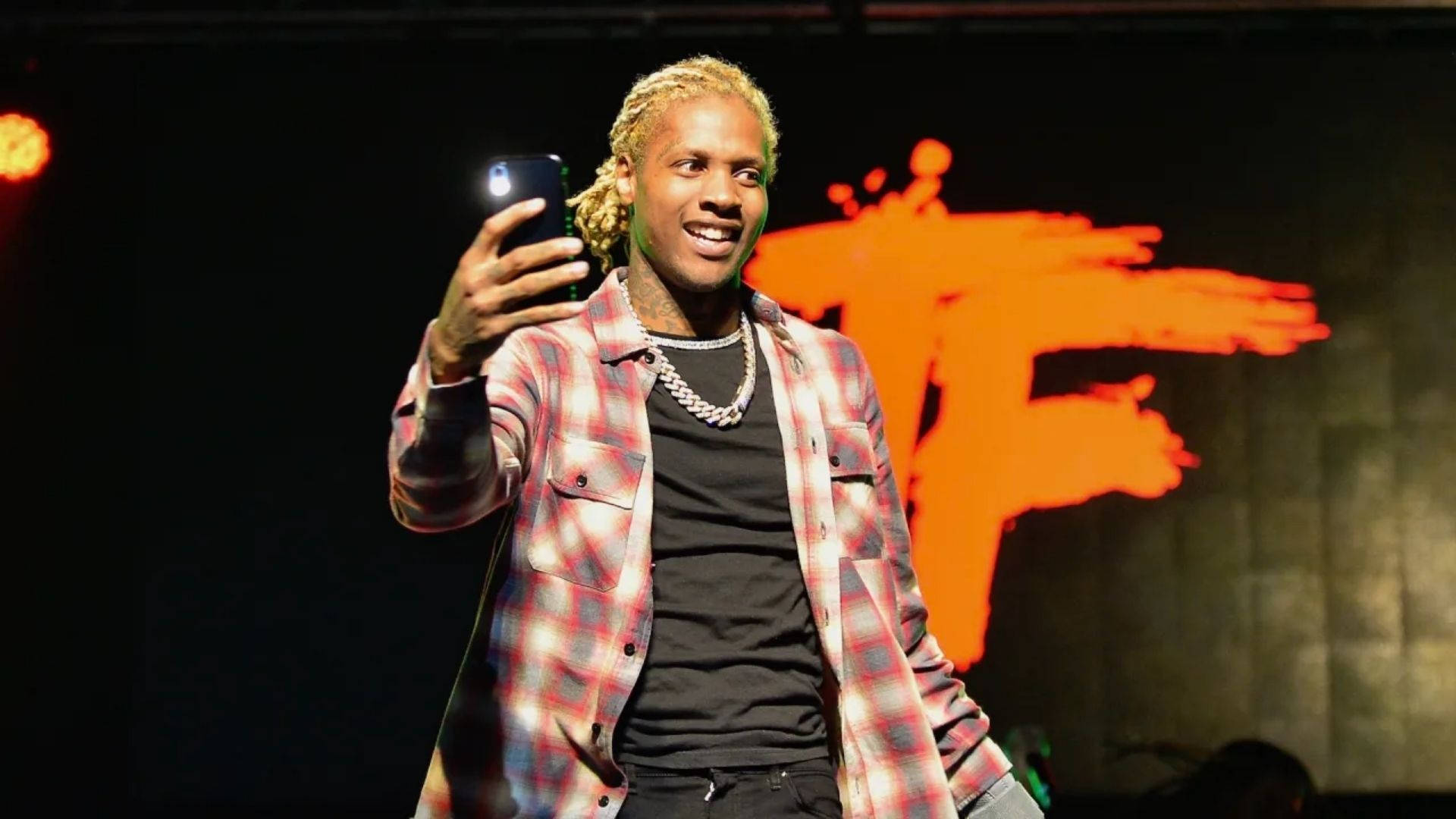 Lil Durk 桌布 壁紙 — 一位微笑的金髮辮子男在明亮的橙色燈光下於舞台上自拍。