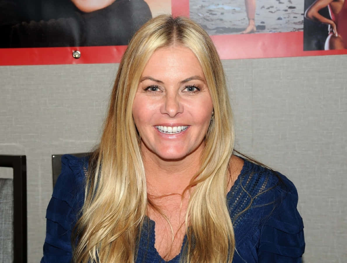 Nicole Eggert 桌布 壁紙 — 一位微笑的金髮長髮女性穿著藍色上衣，背景是貼有海報的灰色牆壁。