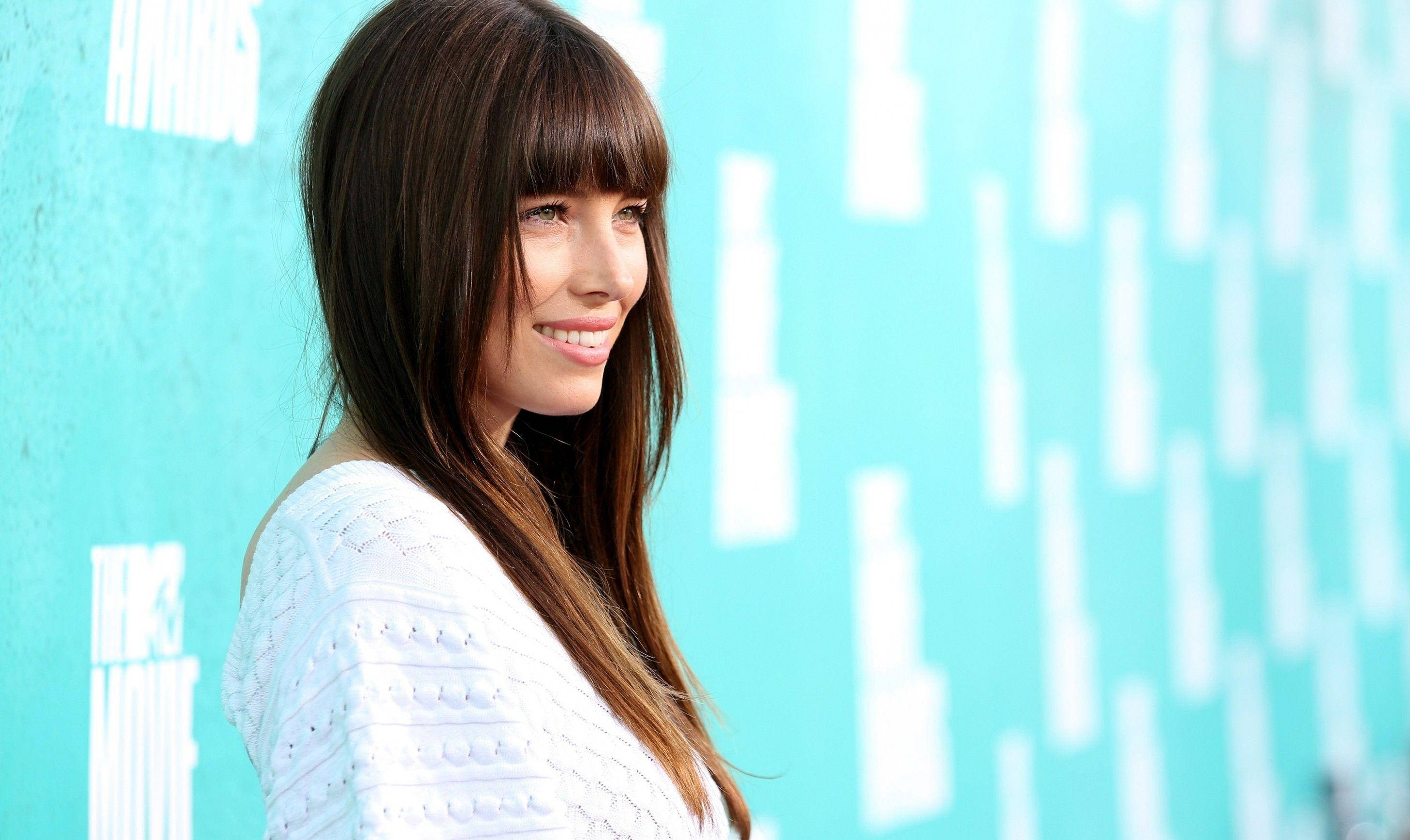 jessica biel 桌布 壁紙 — 一位微笑的長棕髮及瀏海女性，身穿白色紋理上衣，背景是帶有白色字樣的青綠色牆壁。