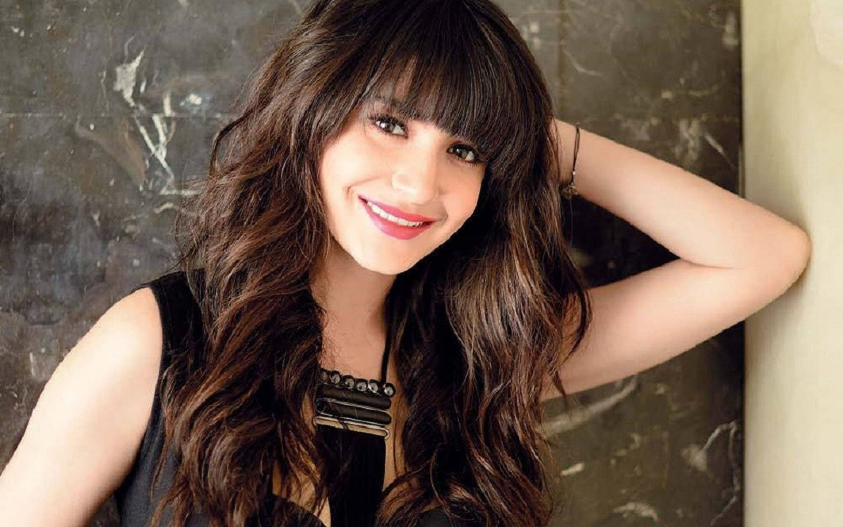 Anushka Sharma 桌布 壁紙 — 一位微笑的長波浪棕色頭髮女性，穿著黑色上衣，靠著深色大理石牆。
