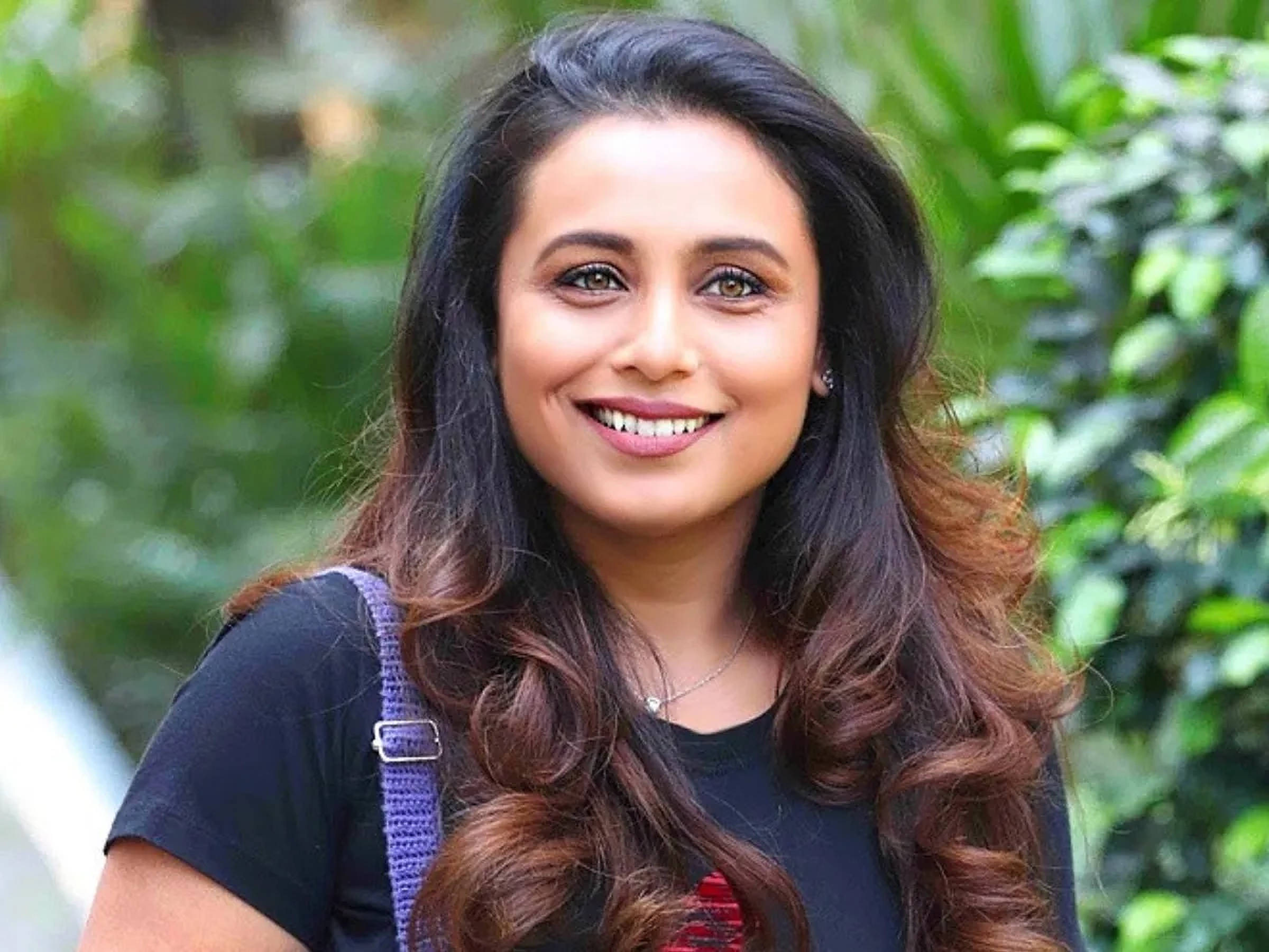 Rani Mukerji 桌布 壁紙 — 一位微笑的長波浪棕色頭髮女性站在戶外，背景是茂密的綠色樹葉。