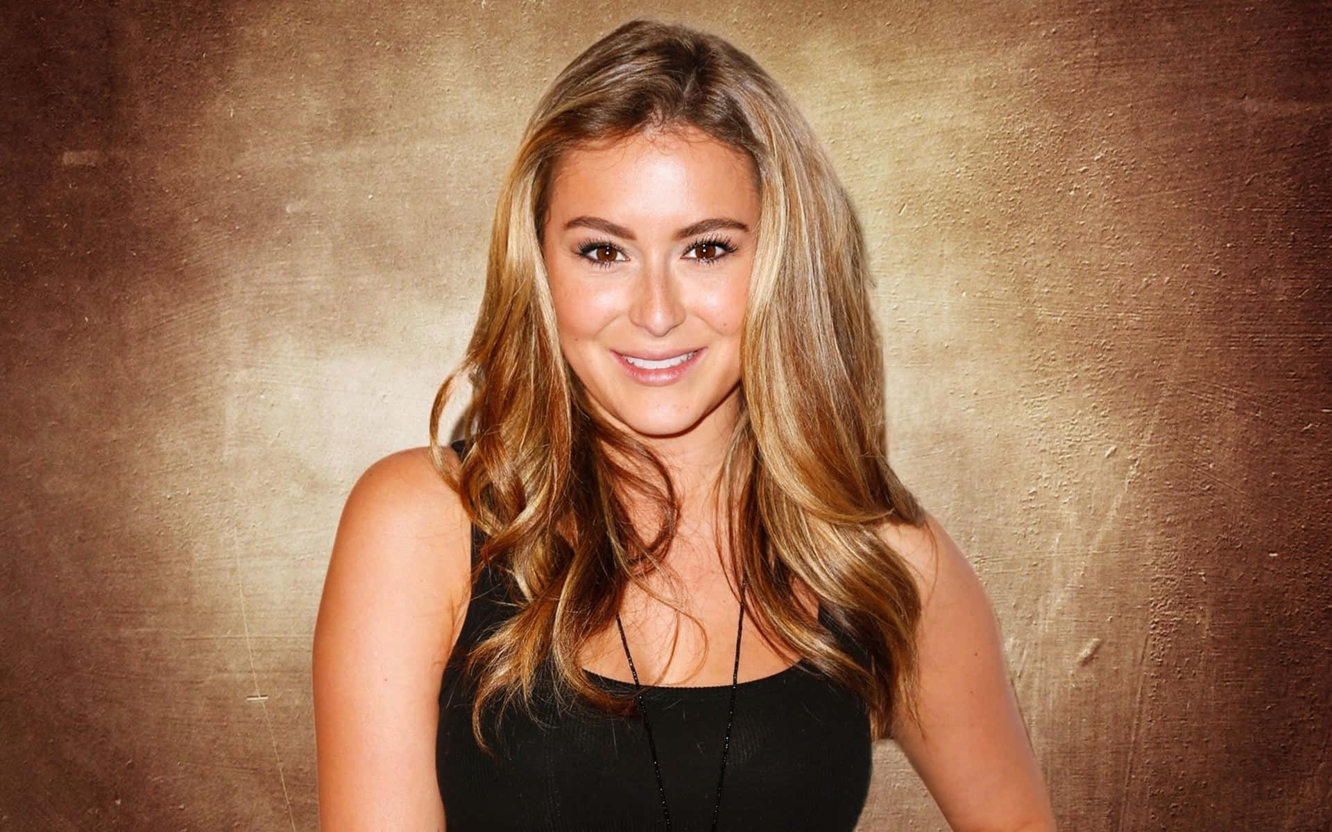 Alexa Penavega 桌布 壁紙 — 一位微笑的長金髮女性穿著黑色上衣，背景是帶有紋理的金色。