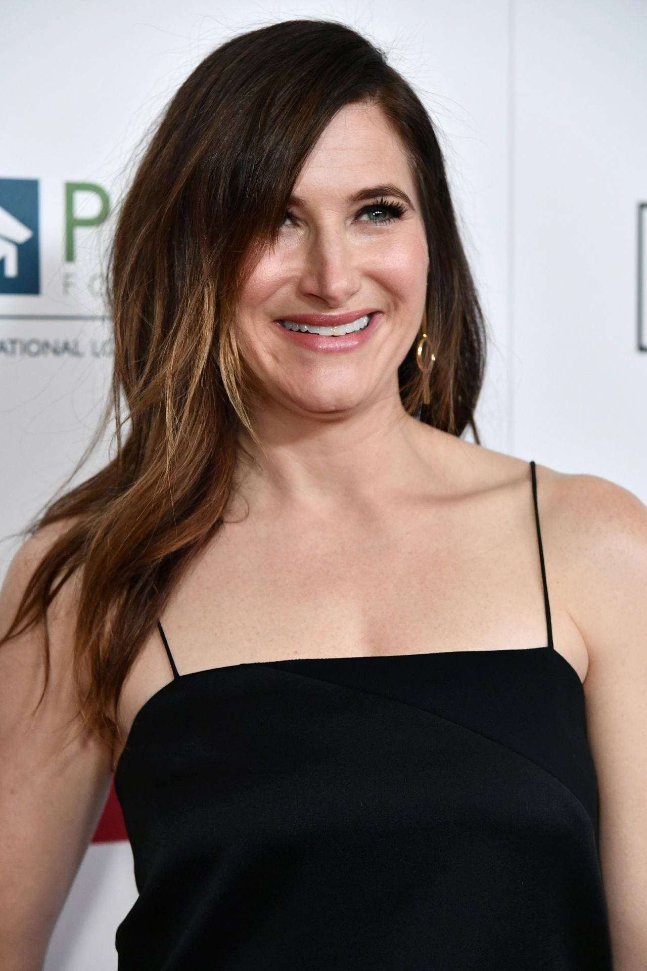 Celebrity 桌布 壁紙 — 一位微笑的長髮棕髮女性，在白色背景的活動中穿著黑色細肩帶洋裝。