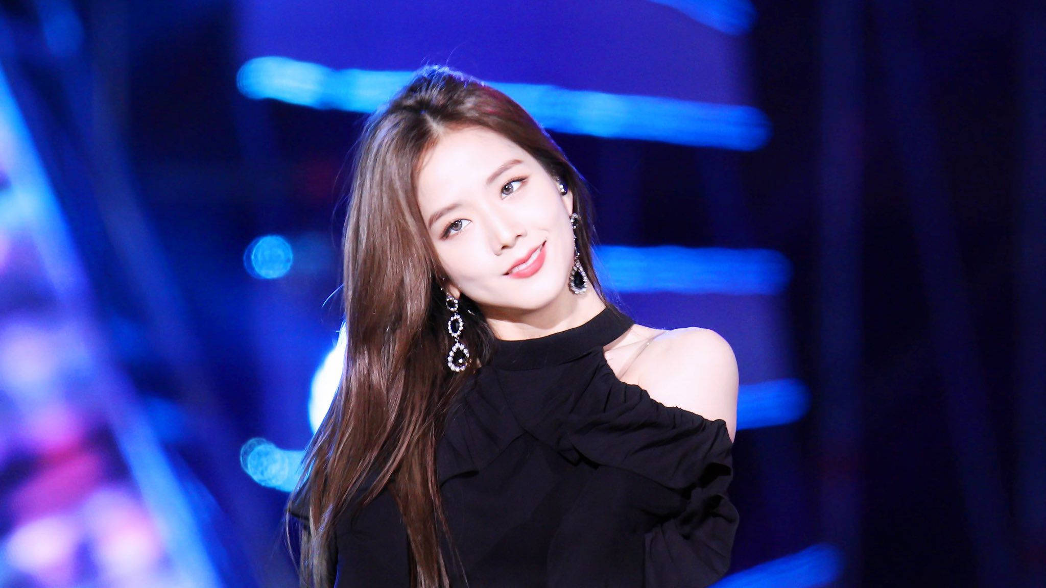 Jisoo Cute 桌布 壁紙 — 一位微笑的長髮棕髮女性，穿著黑色露肩上衣和垂墜耳環，背景是充滿活力的藍色舞台。