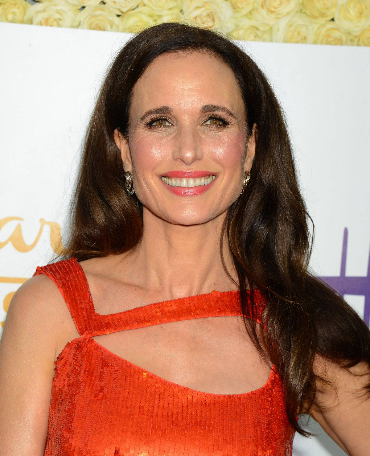 andie macdowell 桌布 壁紙 — 一位微笑的長髮棕髮女性，身穿鮮豔的橙色亮片連衣裙，背景是黃玫瑰和活動標誌。