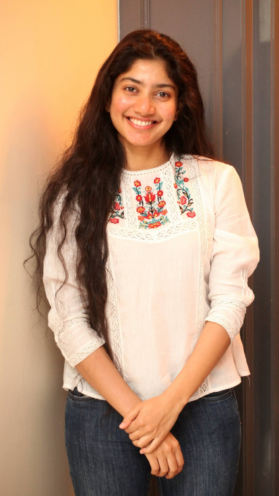 Sai Pallavi 桌布 壁紙 — 一位微笑的長髮深色女子穿著白色刺繡襯衫和牛仔褲，靠著暖色調的室內牆壁。