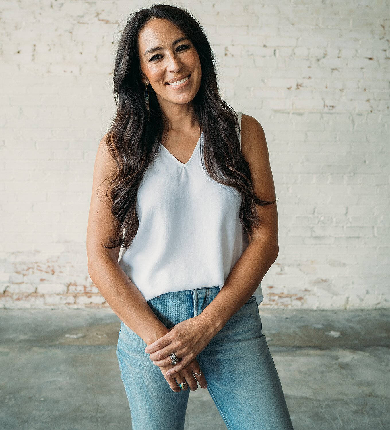 Joanna 桌布 壁紙 — 一位微笑的長髮深色女性自信地站在白色磚牆前，身穿白色上衣和藍色牛仔褲。