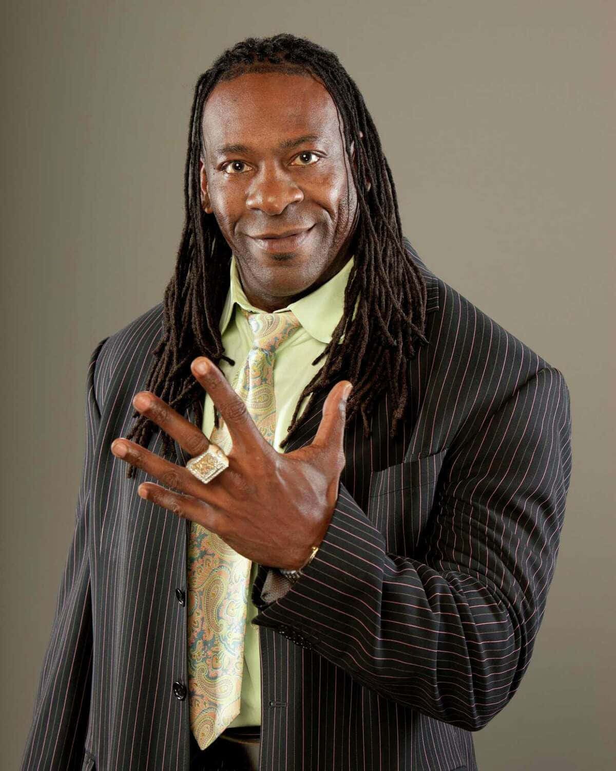 WWE 桌布 壁紙 — 一位微笑的長髮辮男子身穿細條紋西裝和圖案領帶，用戴著大戒指的手做手勢。