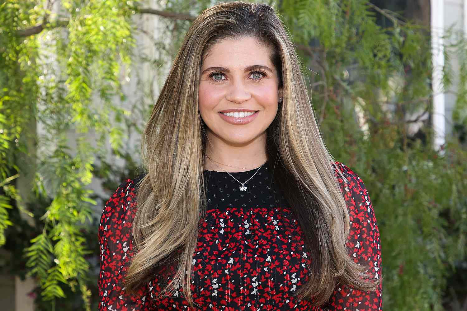 Danielle Fishel 桌布 壁紙 — 一位微笑的長髮金髮女子穿著紅色花卉連衣裙，背景是茂密的綠色樹葉。