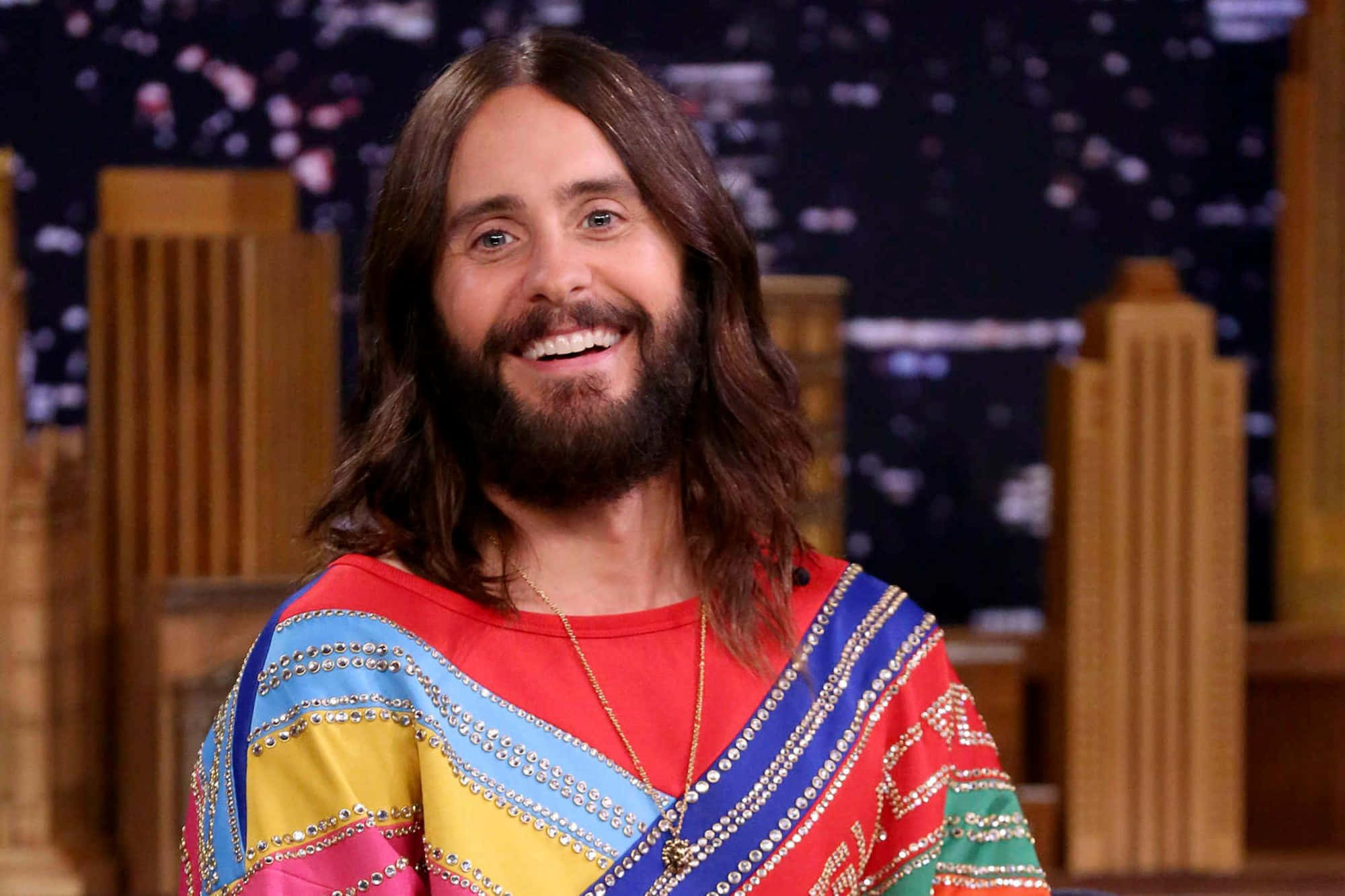 jared leto 桌布 壁紙 — 一位微笑的長髮鬍鬚男，穿著彩色珠飾上衣，背景是城市天際線。
