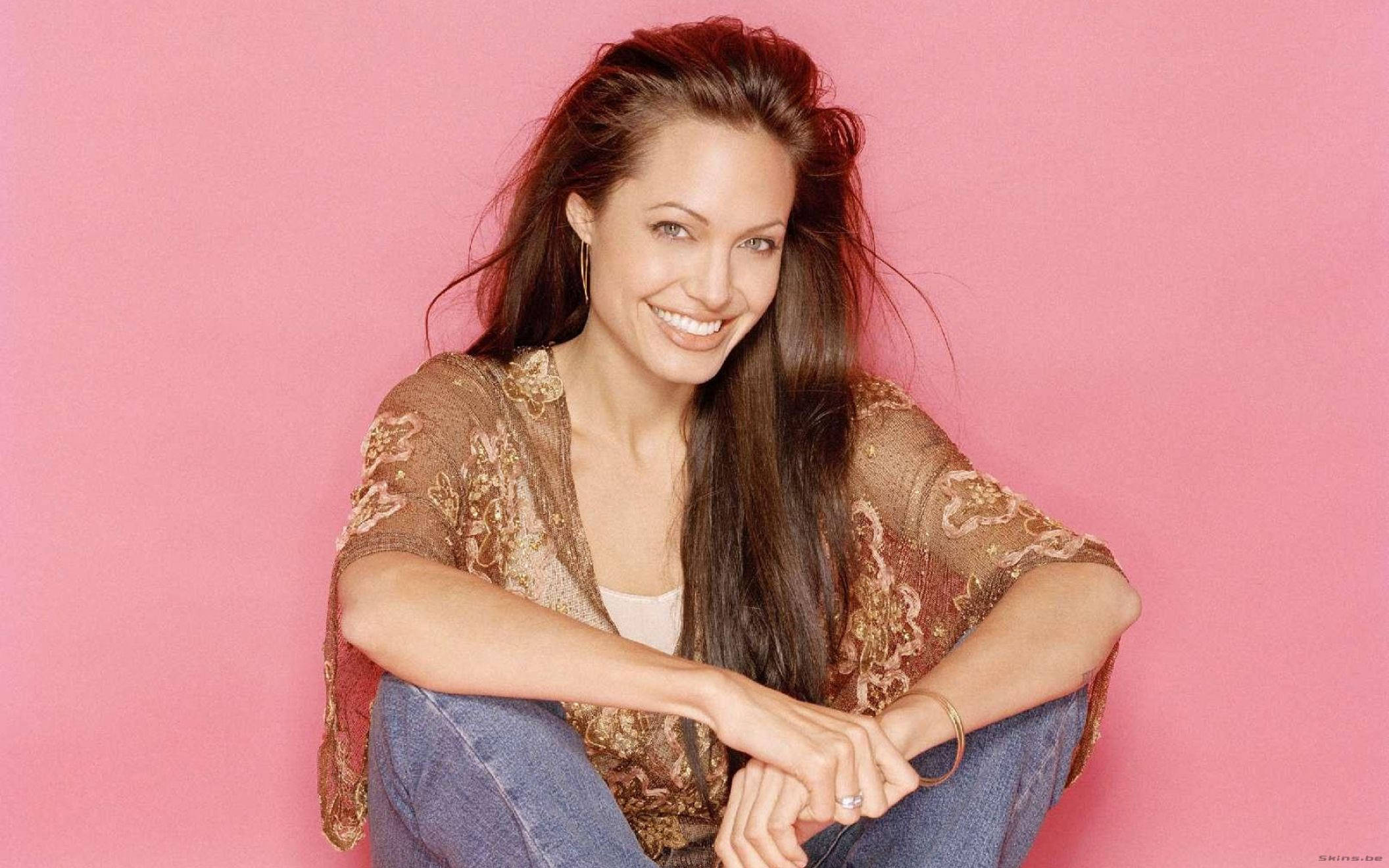 Angelina 桌布 壁紙 — 一位微笑的長黑髮女性，穿著圖案上衣和牛仔褲，隨意地坐在純粉色背景前。