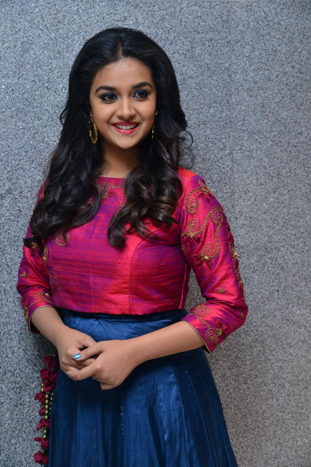 Keerthi Suresh HD 桌布 壁紙 — 一位微笑的長黑髮女性，身穿鮮豔的粉紅色刺繡上衣和藍色裙子，背景是灰色紋理牆壁。