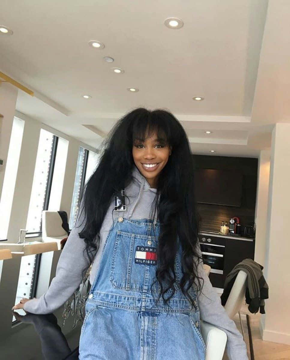 Celebrity 桌布 壁紙 — 一位微笑的長黑髮女性在現代明亮的廚房裡，穿著灰色連帽衫外搭牛仔吊帶褲。