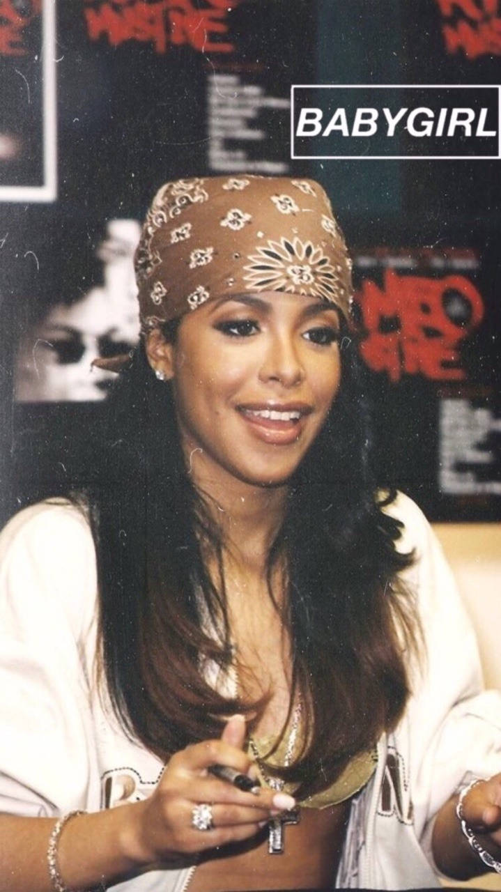 aaliyah 桌布 壁紙 — 一位微笑的長黑髮女性戴著圖案頭巾和白色上衣，在光線昏暗的塗鴉牆房間裡拿著筆。