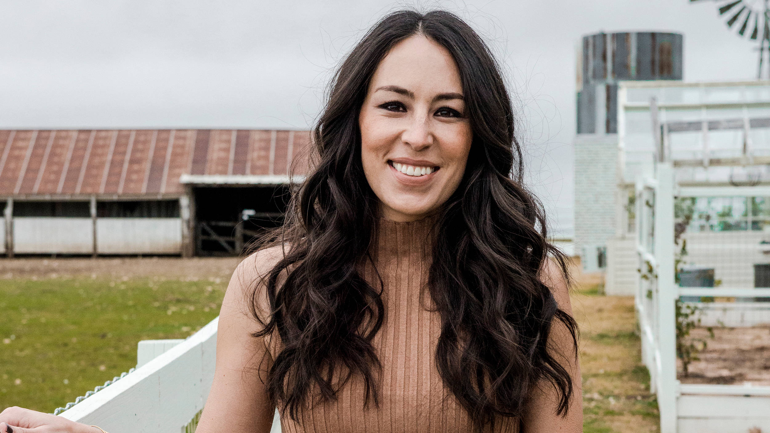 Joanna 桌布 壁紙 — 一位微笑的長黑髮女性站在戶外白色圍欄附近，背景有穀倉和風車，天空多雲。
