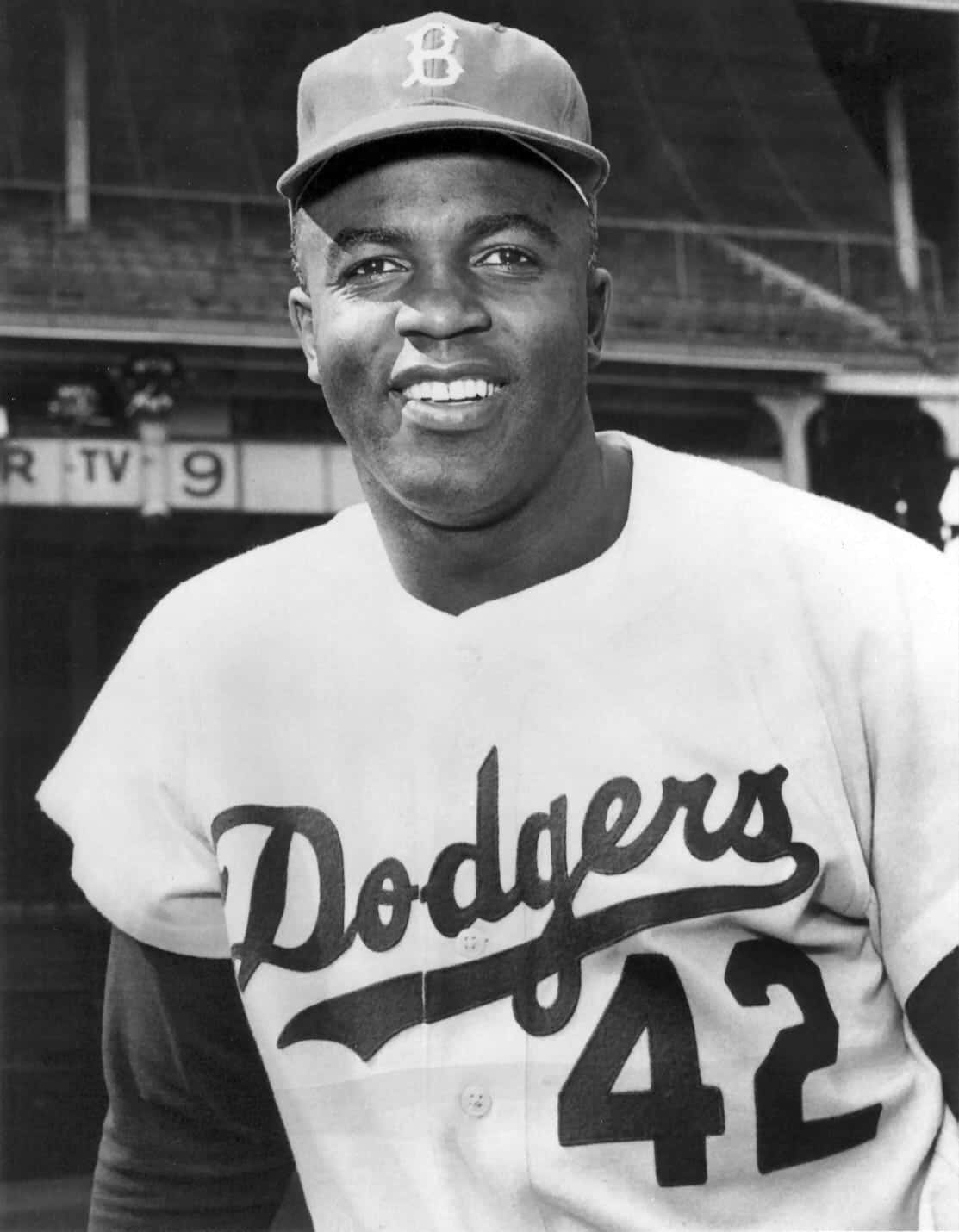 Jackie Robinson 桌布 壁紙 — 一位微笑的非裔棒球選手身穿道奇隊球衣，背號42，戴著球帽，自信地站在體育場的球員休息區。
