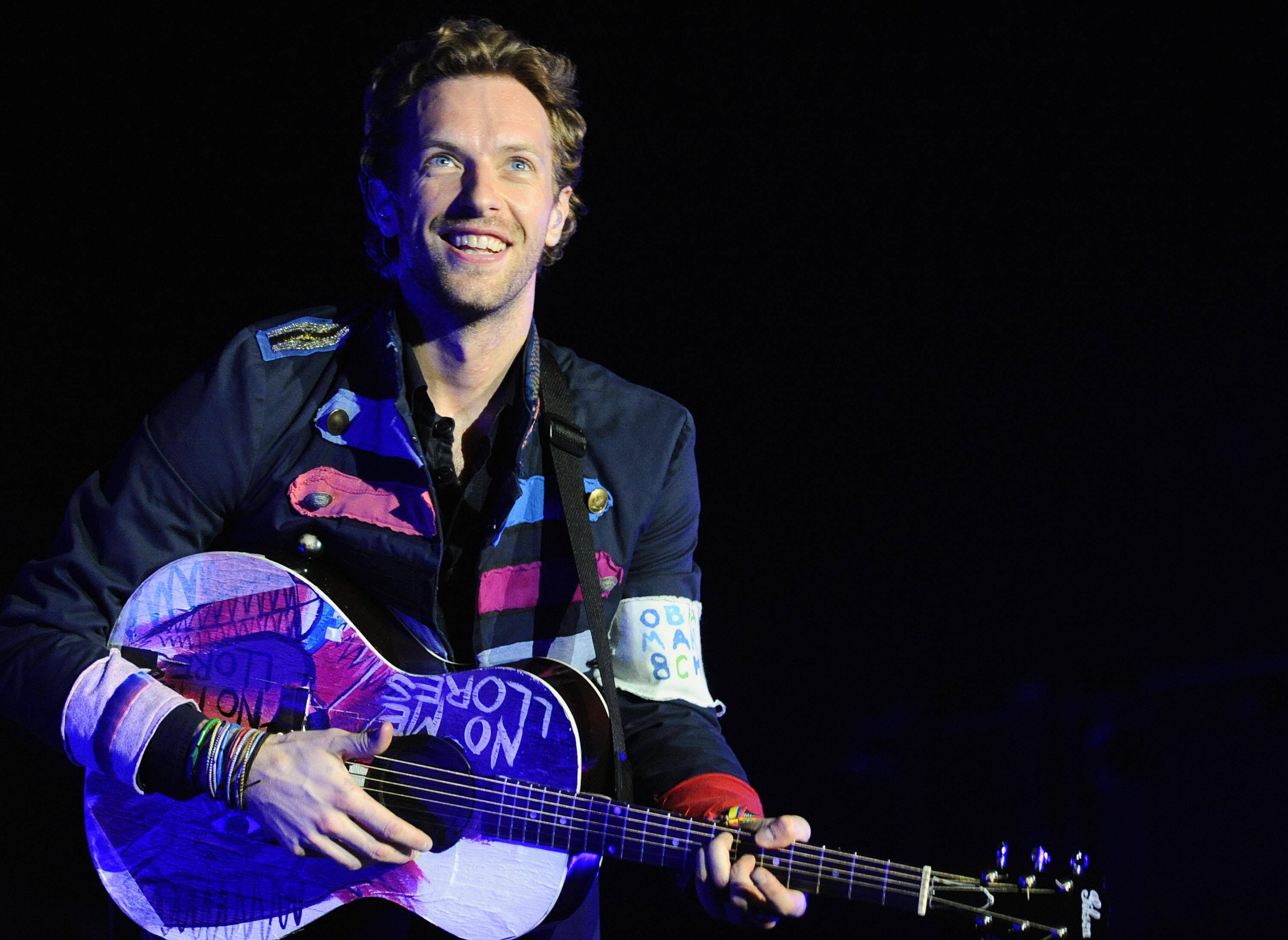 Chris Martin 桌布 壁紙 — 一位微笑的音樂家在黑色背景的舞台燈光下彈奏色彩鮮豔的原聲吉他。