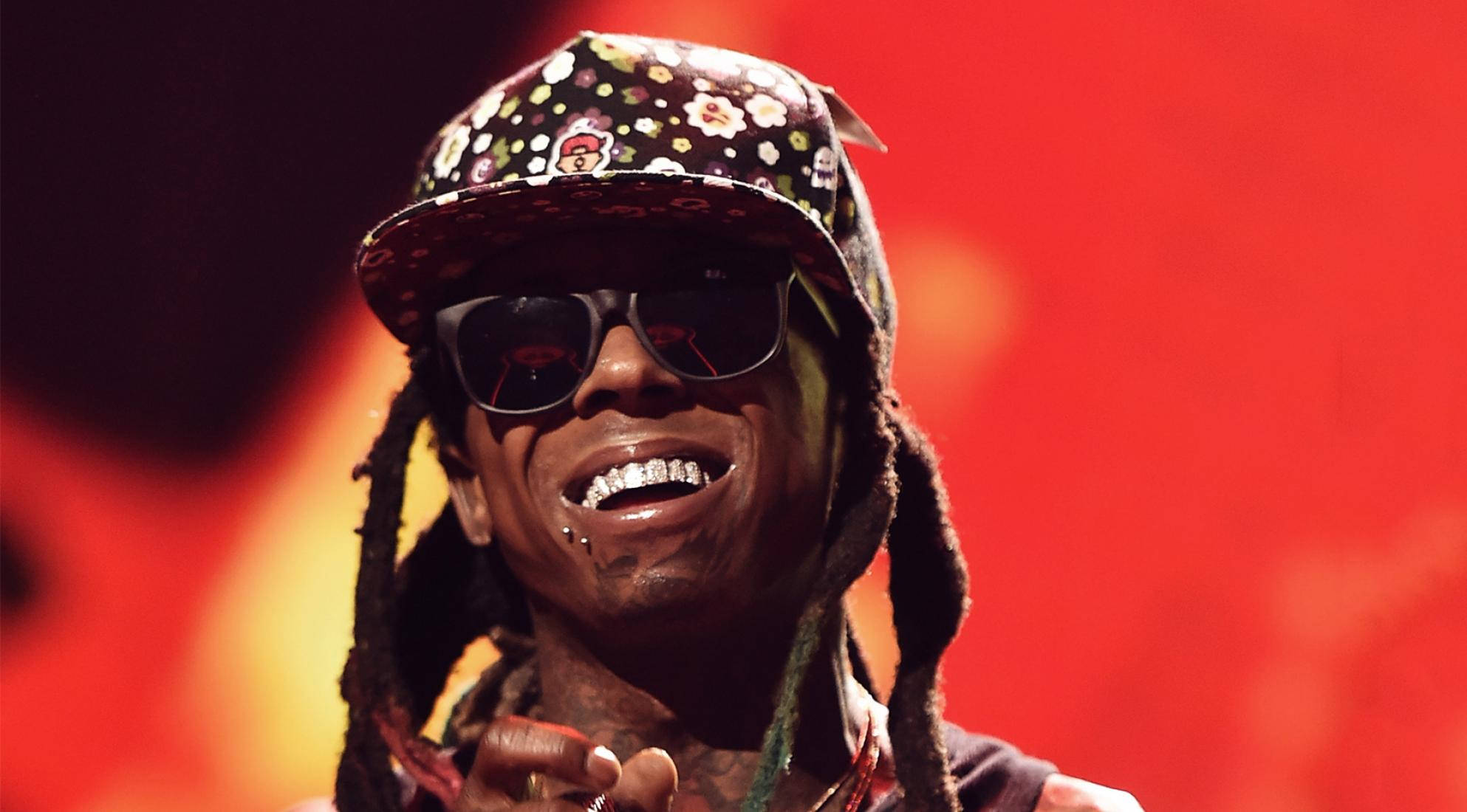 Lil Wayne 桌布 壁紙 — 一位微笑的饒舌歌手，留著辮子頭，戴著花卉圖案帽子，在舞台燈光下對著火紅背景咧嘴笑。