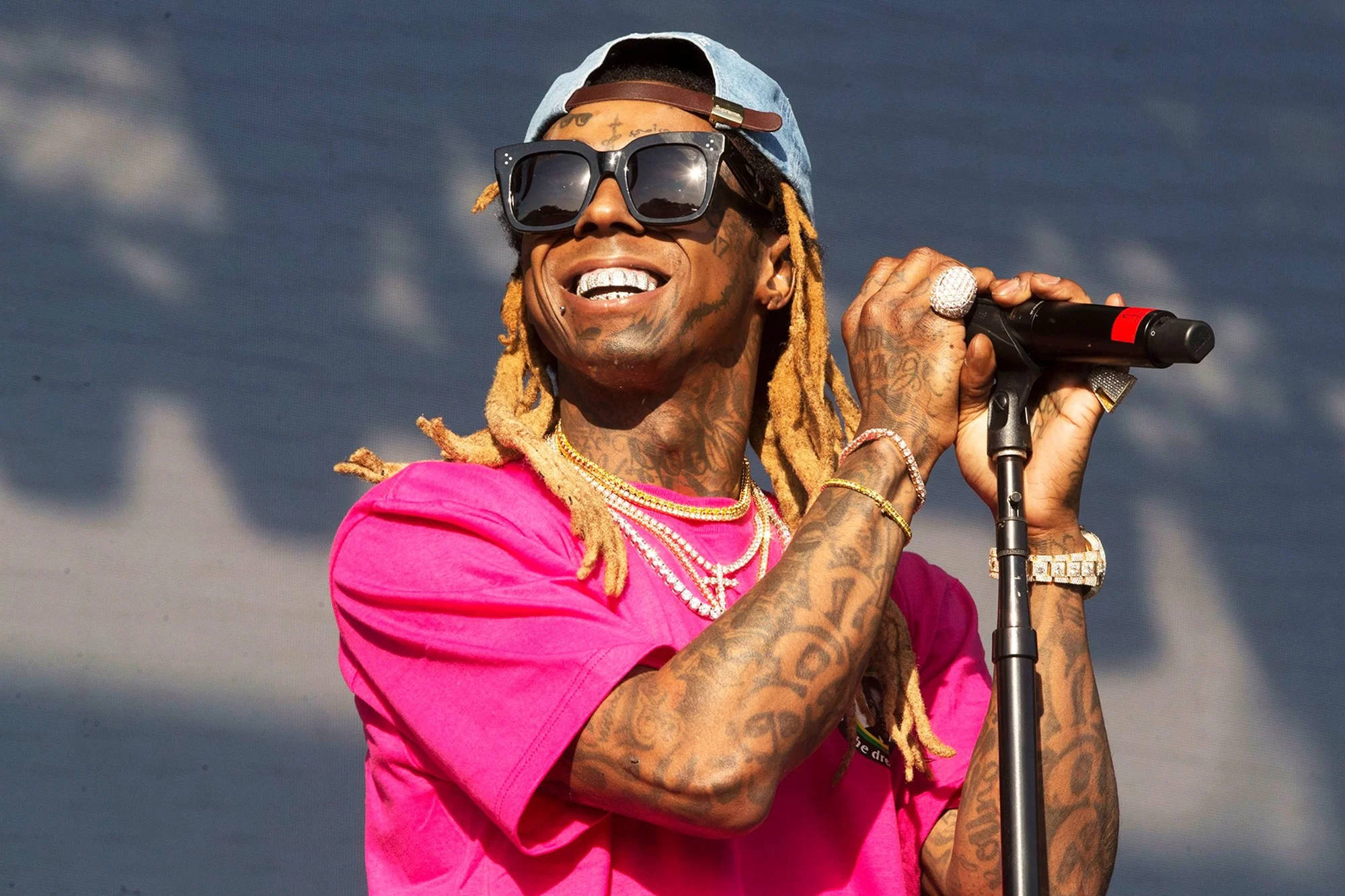 Lil Wayne 桌布 壁紙 — 一位微笑的饒舌歌手，留著辮子頭和刺青，穿著亮粉色襯衫和太陽眼鏡對著麥克風表演。