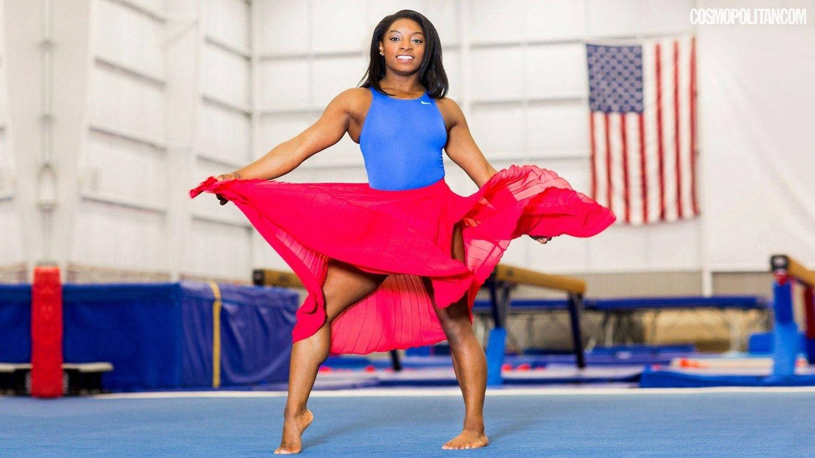 Simone Biles 桌布 壁紙 — 一位微笑的體操運動員身穿藍色緊身衣和飄逸的紅色裙子，在藍色墊子上以美國國旗為背景動感地擺出姿勢。