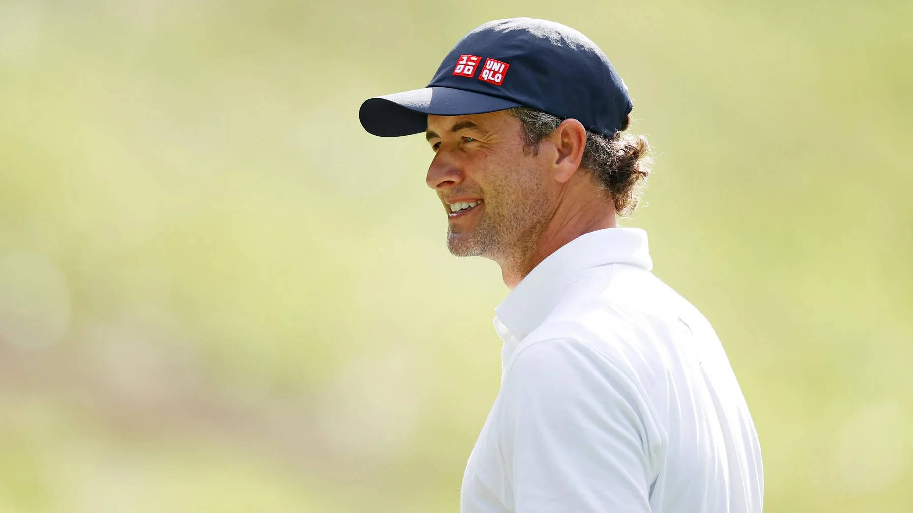 adam scott 桌布 壁紙 — 一位微笑的高爾夫球手，身穿白色Polo衫和深藍色帽子，站在柔和的綠色模糊背景前。