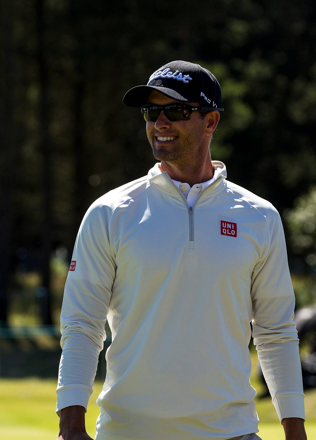 adam scott 桌布 壁紙 — 一位微笑的高爾夫球手，身穿白色長袖襯衫和黑色帽子，站在陽光明媚的高爾夫球場上，背景有樹木。
