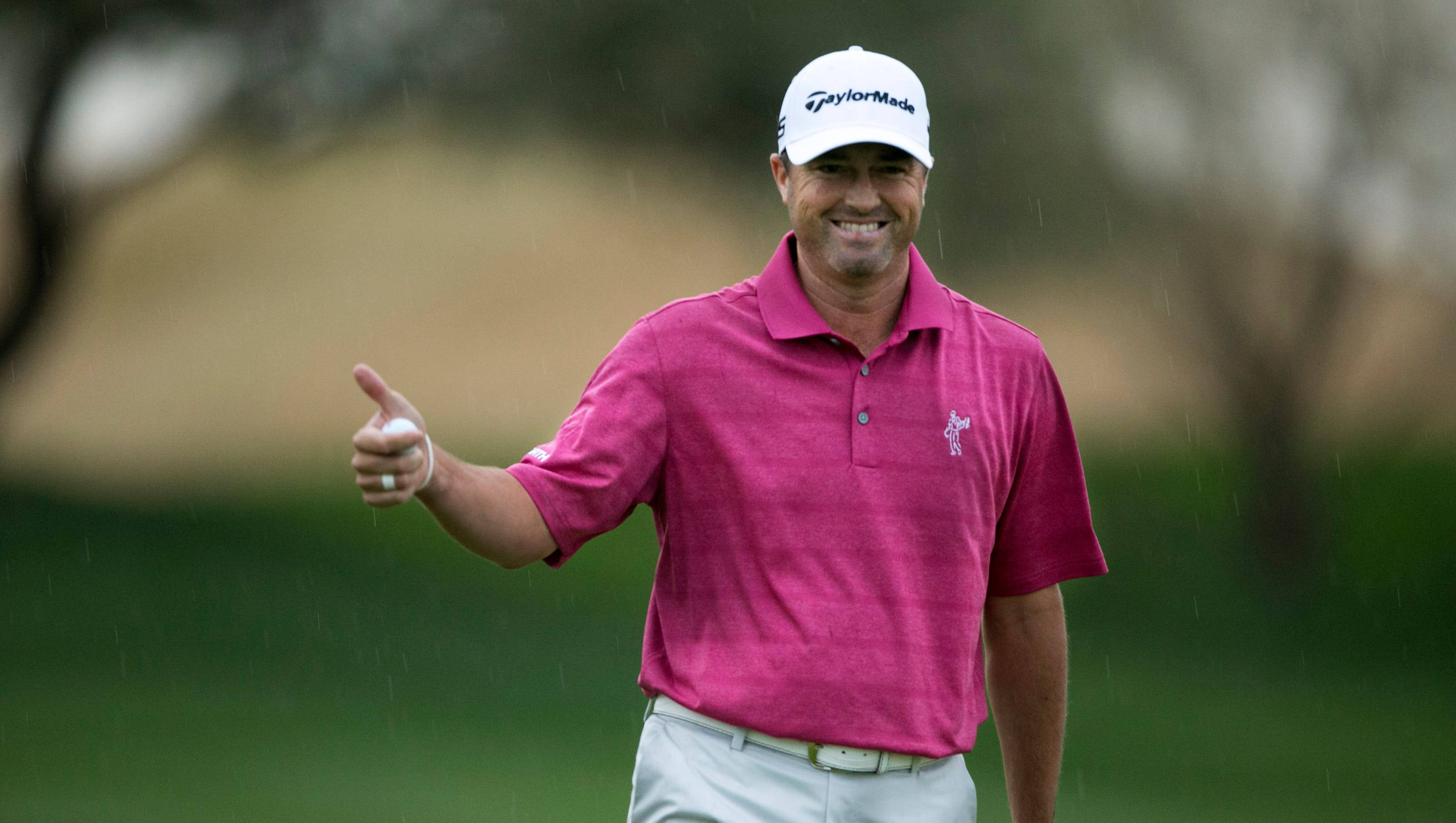 Ryan Palmer 桌布 壁紙 — 一位微笑的高爾夫球手，身穿粉紅色Polo衫和白色帽子，在雨中高爾夫球場上行走時豎起大拇指。