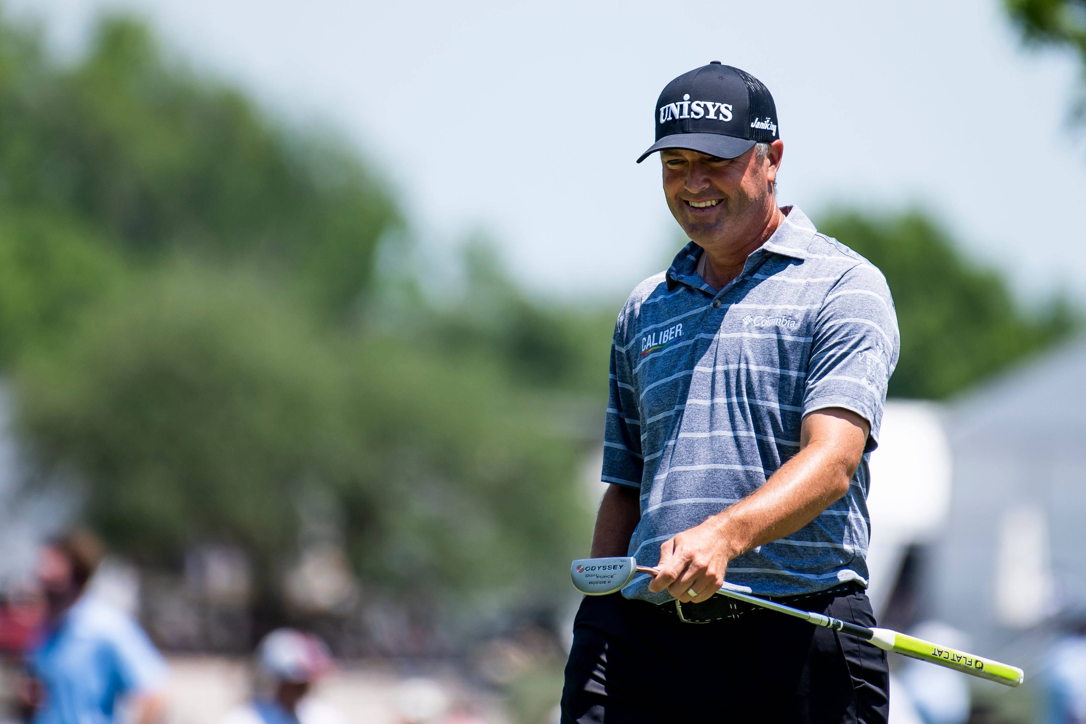 Ryan Palmer 桌布 壁紙 — 一位微笑的高爾夫球手，身穿藍色條紋襯衫和帽子，在陽光明媚的球場上拿著推桿，身後是模糊的綠色樹木。