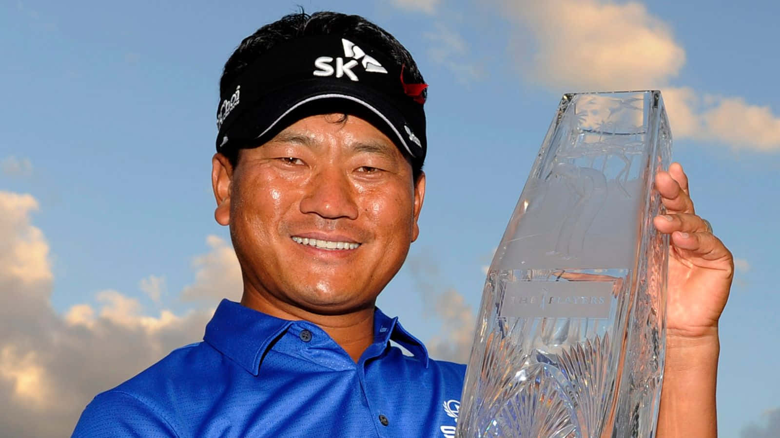Trophy 桌布 壁紙 — 一位微笑的高爾夫球手，身穿藍色襯衫和黑色帽子，自豪地舉起一個大型水晶獎盃，背景是晴朗的藍天和蓬鬆的白雲。