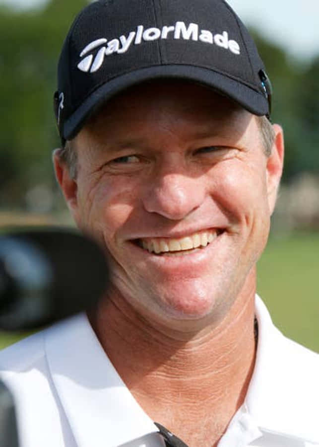 高爾夫 桌布 壁紙 — 一位微笑的高爾夫球手戴著黑色TaylorMade帽子和白色Polo衫，在陽光明媚的日子裡站在戶外，身後是模糊的綠色植物。