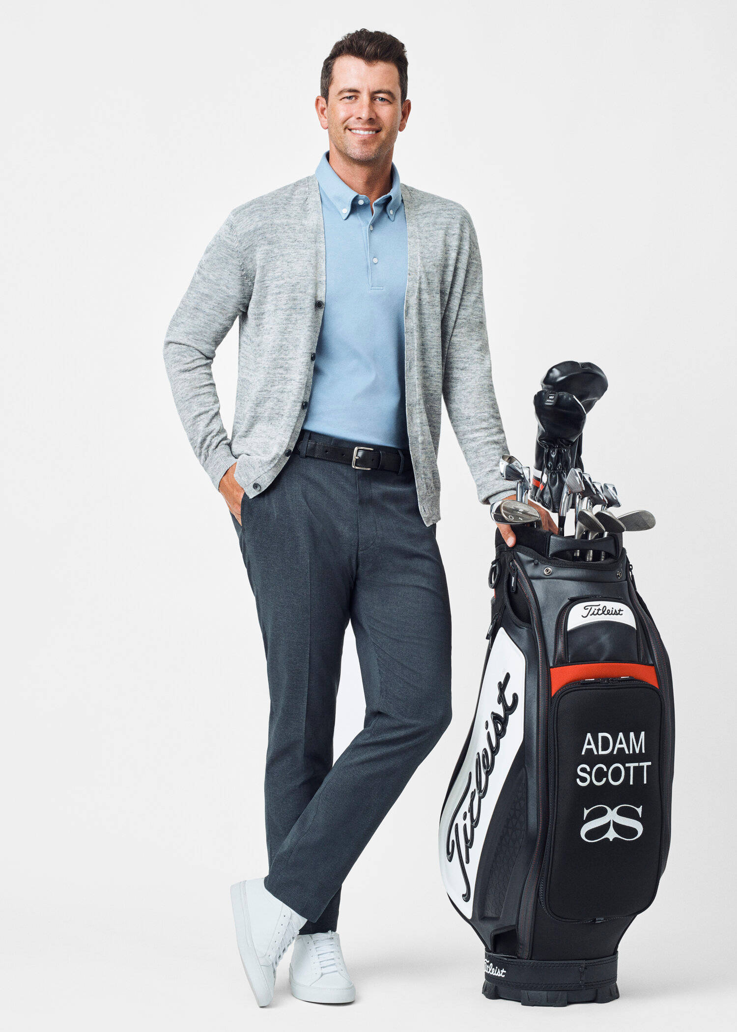 adam scott 桌布 壁紙 — 一位微笑的高爾夫球手穿著淺藍色Polo衫和灰色開襟衫，自信地站在他的品牌高爾夫球袋旁邊，背景是純白色。