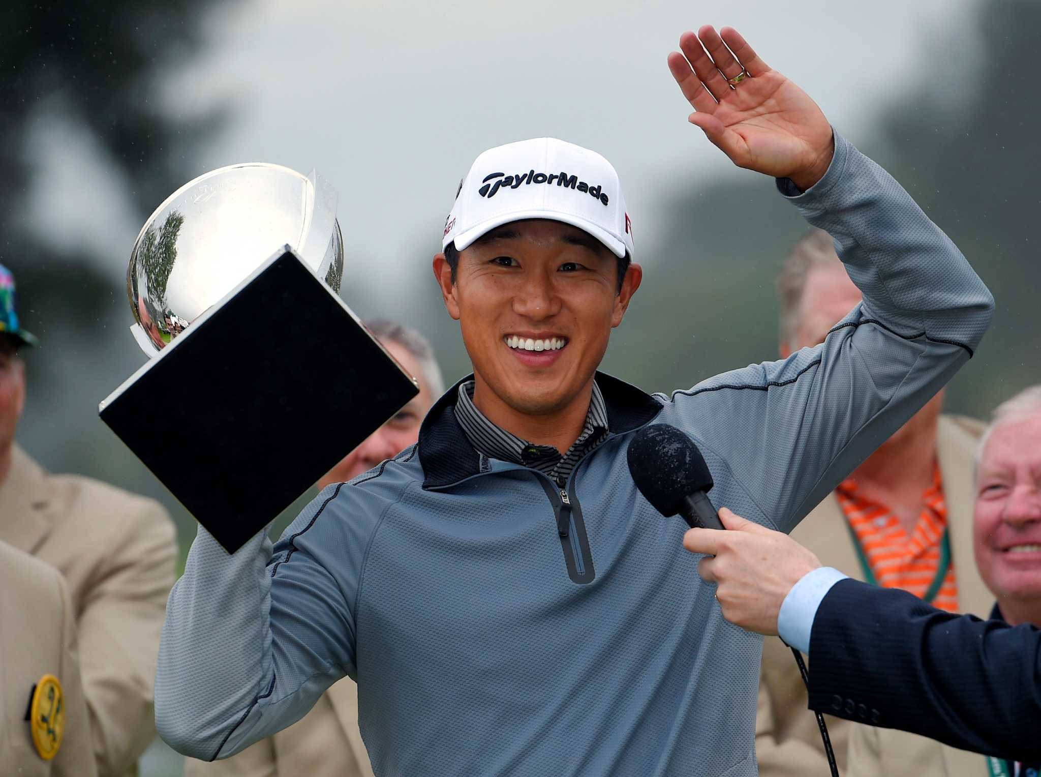 Trophy 桌布 壁紙 — 一位微笑的高爾夫球手穿著灰色套頭衫和白色帽子，在雨中高爾夫錦標賽中揮手向球迷致意，同時手持一個大型銀色獎盃。