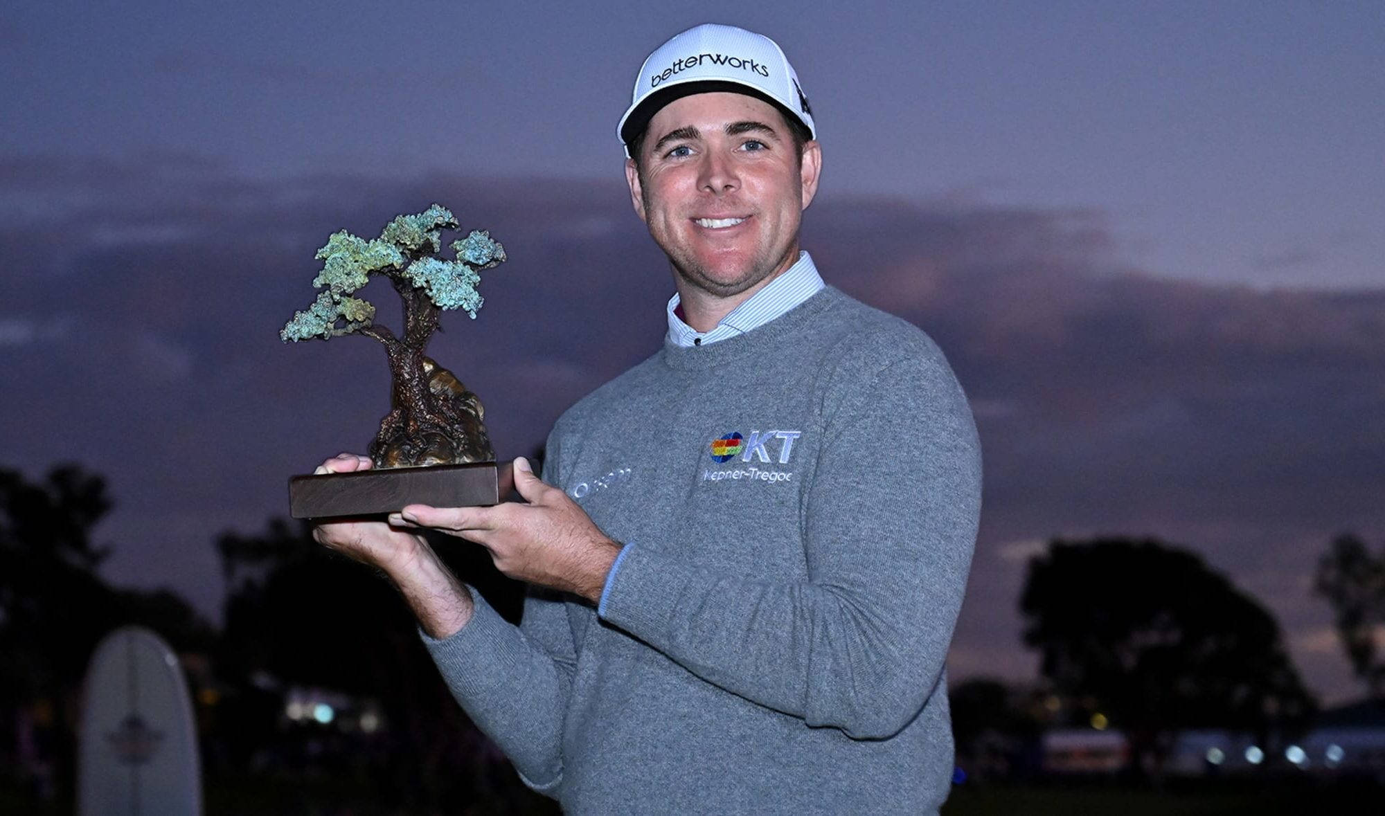 Trophy 桌布 壁紙 — 一位微笑的高爾夫球手穿著灰色毛衣，在暮色天空和柔和紫色雲彩的背景下，手持盆景樹獎盃。