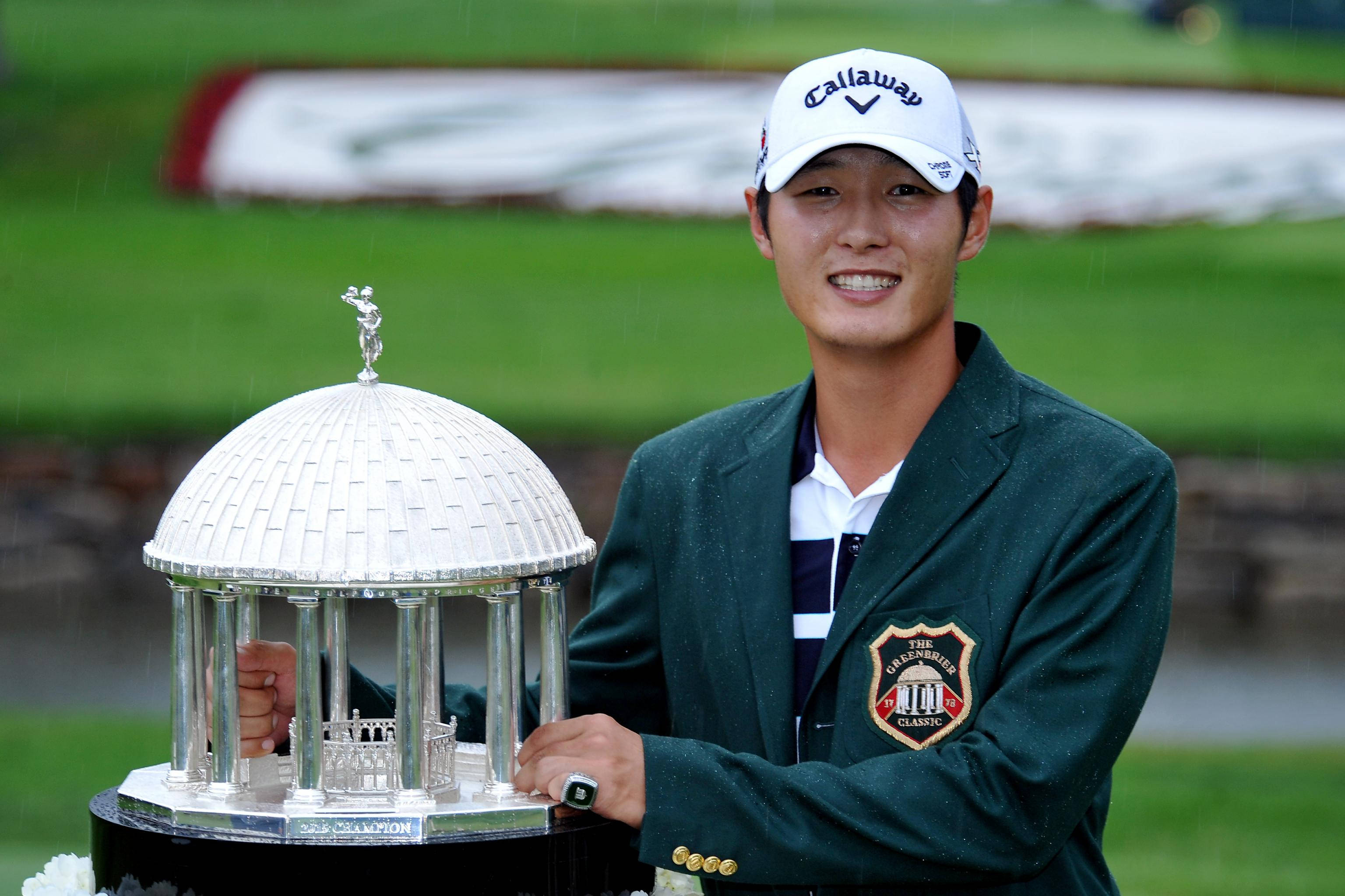 Champion 桌布 壁紙 — 一位微笑的高爾夫球手穿著綠色外套，在潮濕的高爾夫球場上拿著一個大的銀色獎盃。
