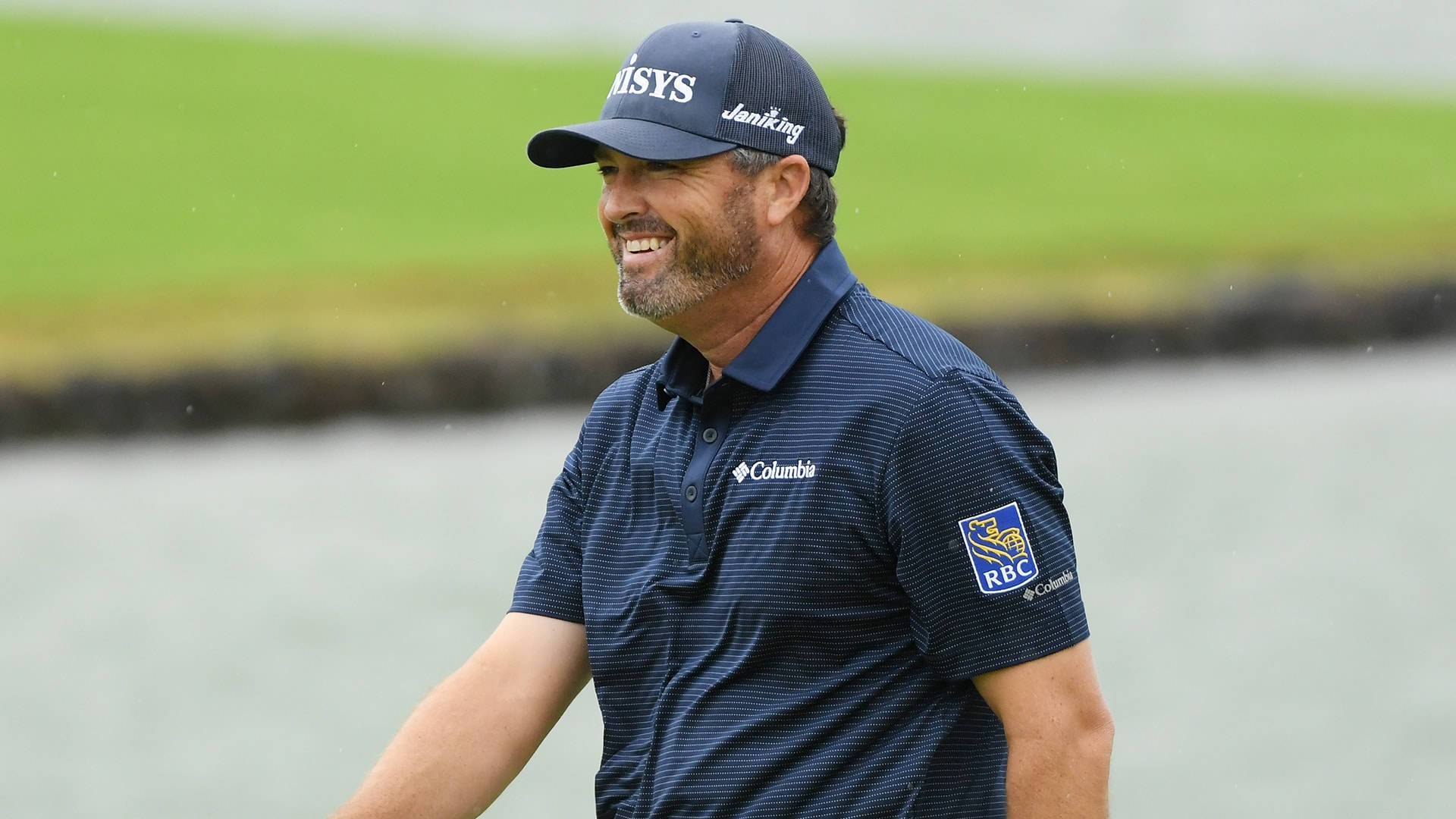 Ryan Palmer 桌布 壁紙 — 一位微笑的高爾夫球手身穿海軍藍Columbia Polo衫和帽子，在翠綠球場的水障礙旁行走。