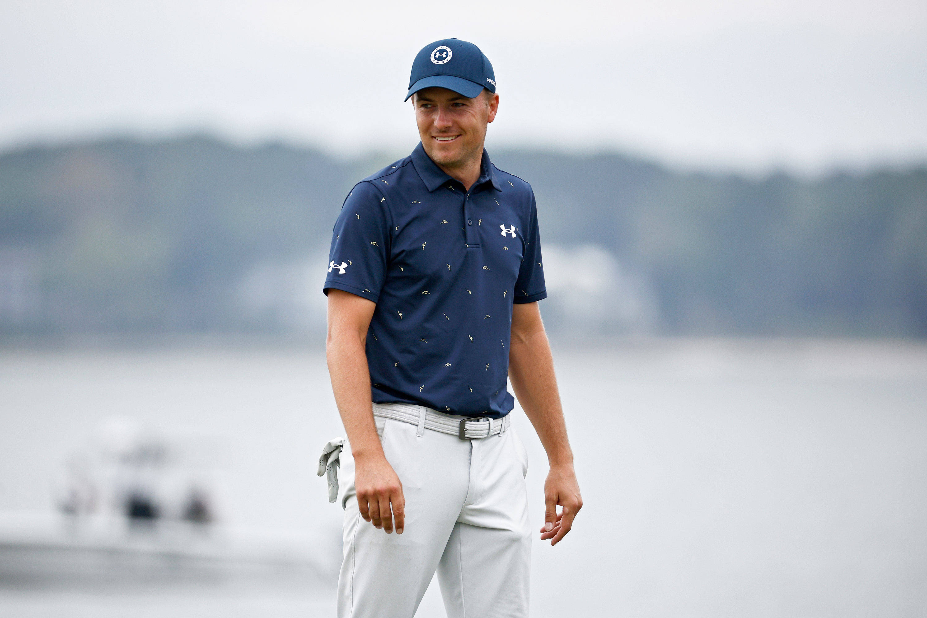 Stylish Boy 桌布 壁紙 — 一位微笑的高爾夫球手身穿海軍藍Polo衫和白色長褲，站在平靜的湖邊，背景是霧氣繚繞的森林山丘。