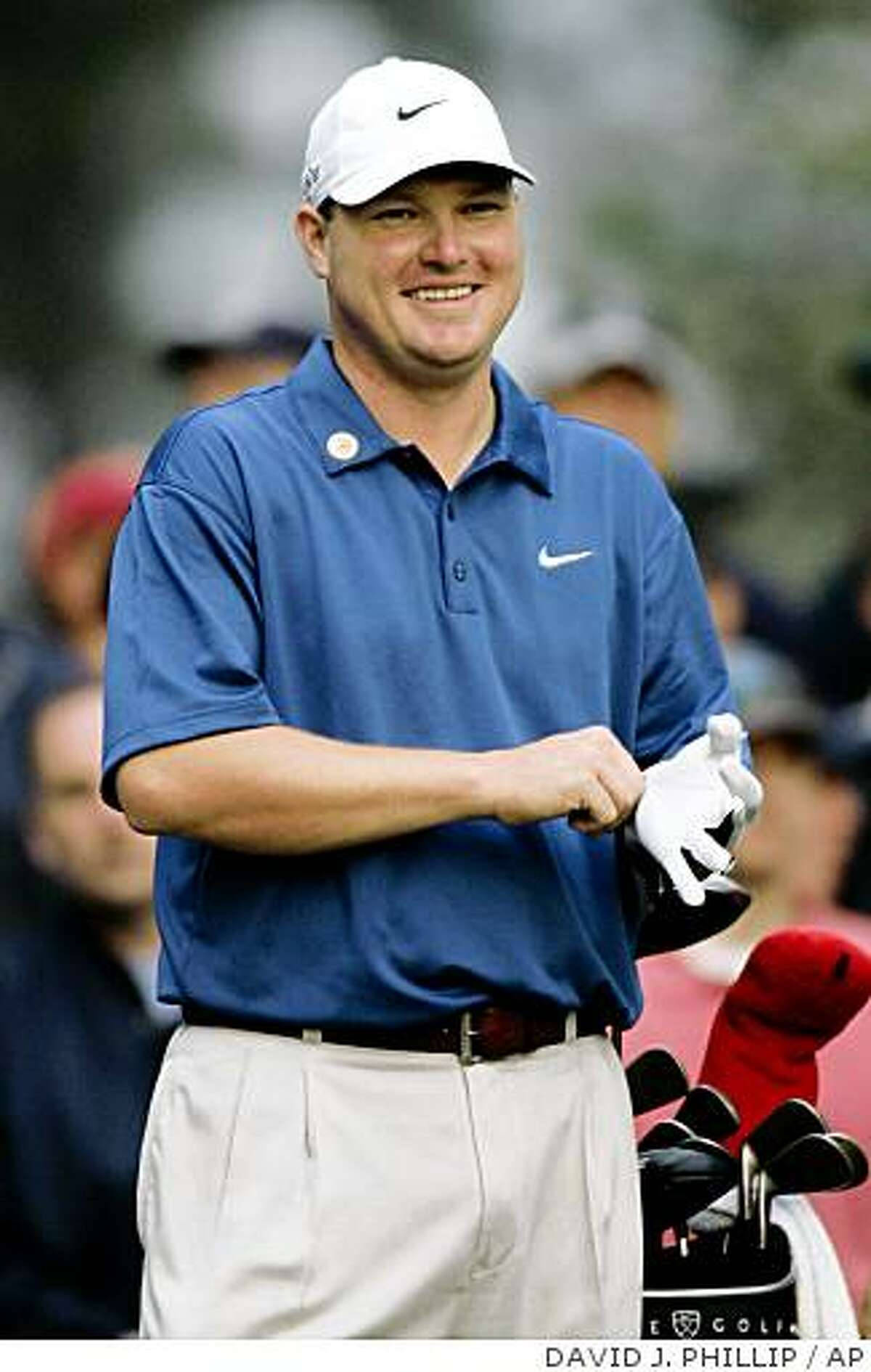 高爾夫 桌布 壁紙 — 一位微笑的高爾夫球手身穿藍色Nike Polo衫和白色帽子，站在高爾夫球袋旁，手持手套。