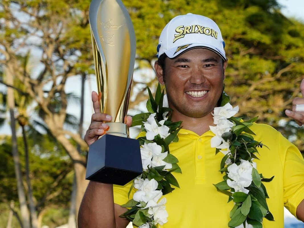 Trophy 桌布 壁紙 — 一位微笑的高爾夫球手身穿黃色襯衫和白色帽子，自豪地舉起金色獎盃，脖子上戴著白色花環，背景是茂密的綠樹。
