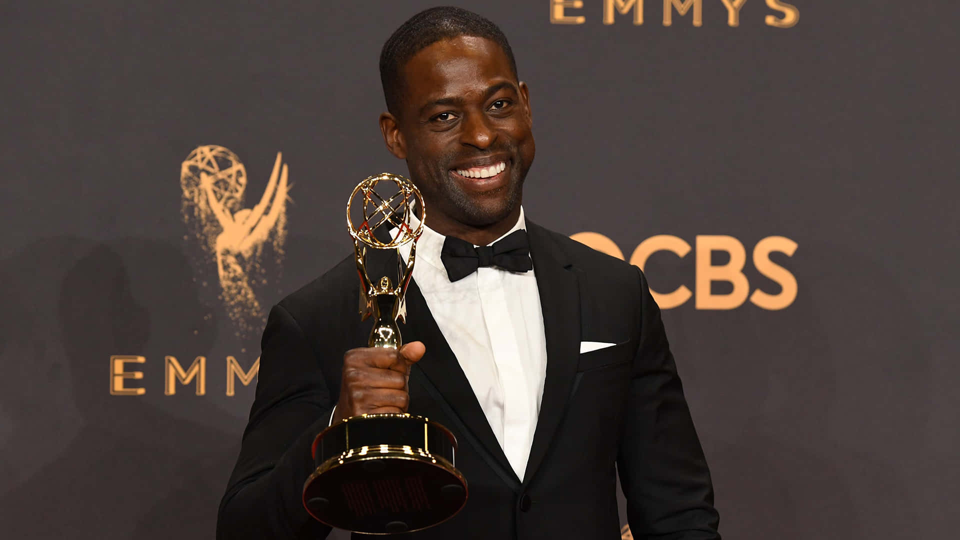Sterling K Brown 桌布 壁紙 — 一位微笑的黑人男士身穿燕尾服，在帶有「EMMYS」和「CBS」標誌的深色背景上自豪地手持金色艾美獎獎座。