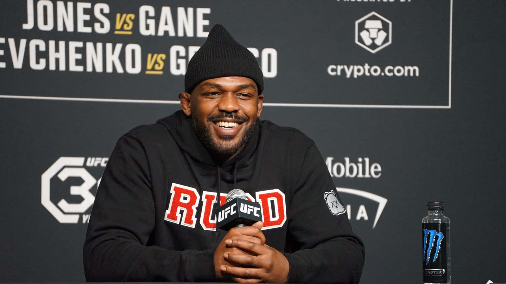 UFC 桌布 壁紙 — 一位微笑的黑人男子戴著黑色無簷便帽和連帽衫，在贊助商標誌後面的新聞活動中對著UFC麥克風講話。