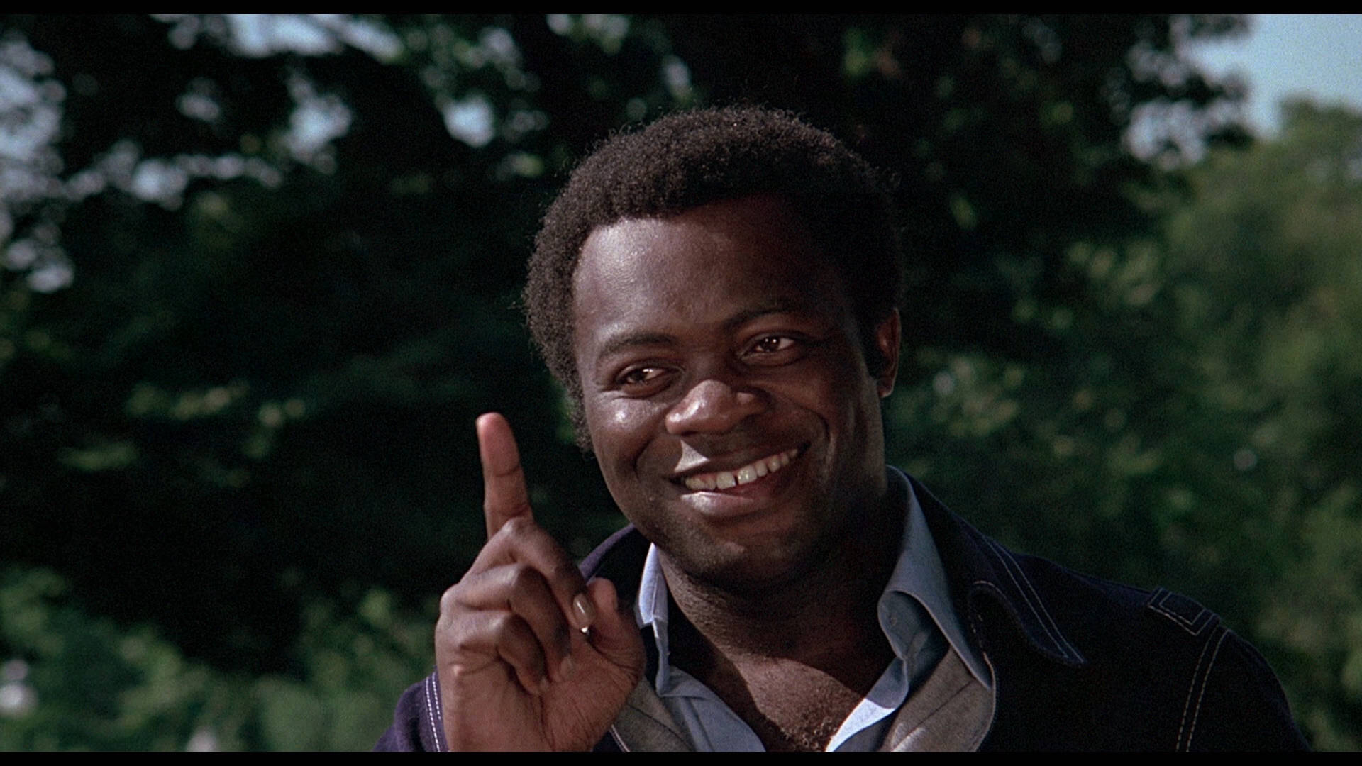 演員 桌布 壁紙 — 一位微笑的黑人男子身穿牛仔外套，右食指向上指，背景是模糊的綠色樹木。