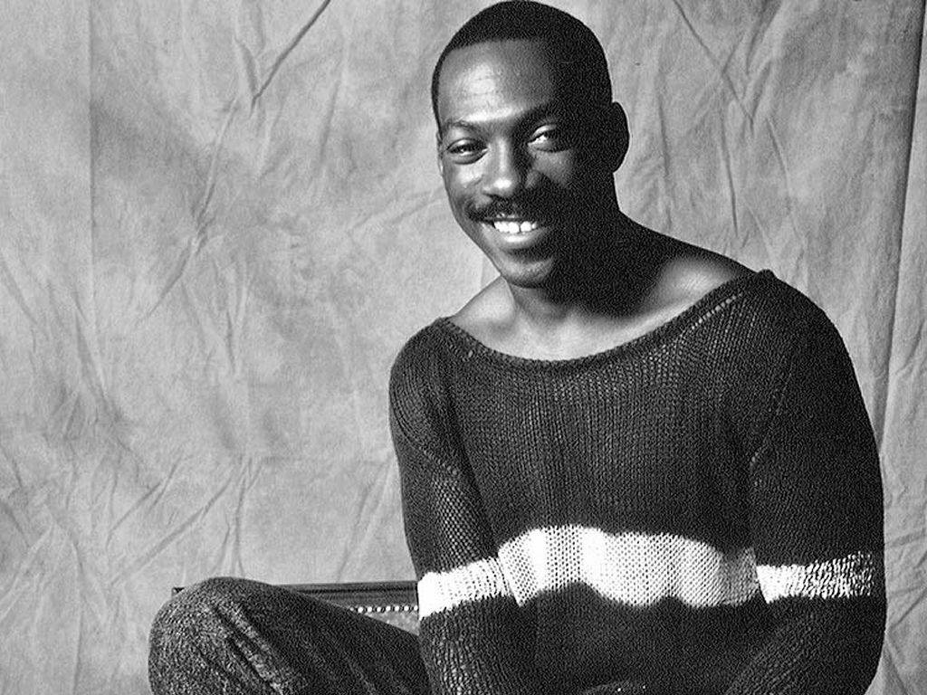 eddie murphy 桌布 壁紙 — 一位微笑的黑人男性穿著針織毛衣，在皺褶的攝影棚背景前擺出經典黑白肖像姿勢。