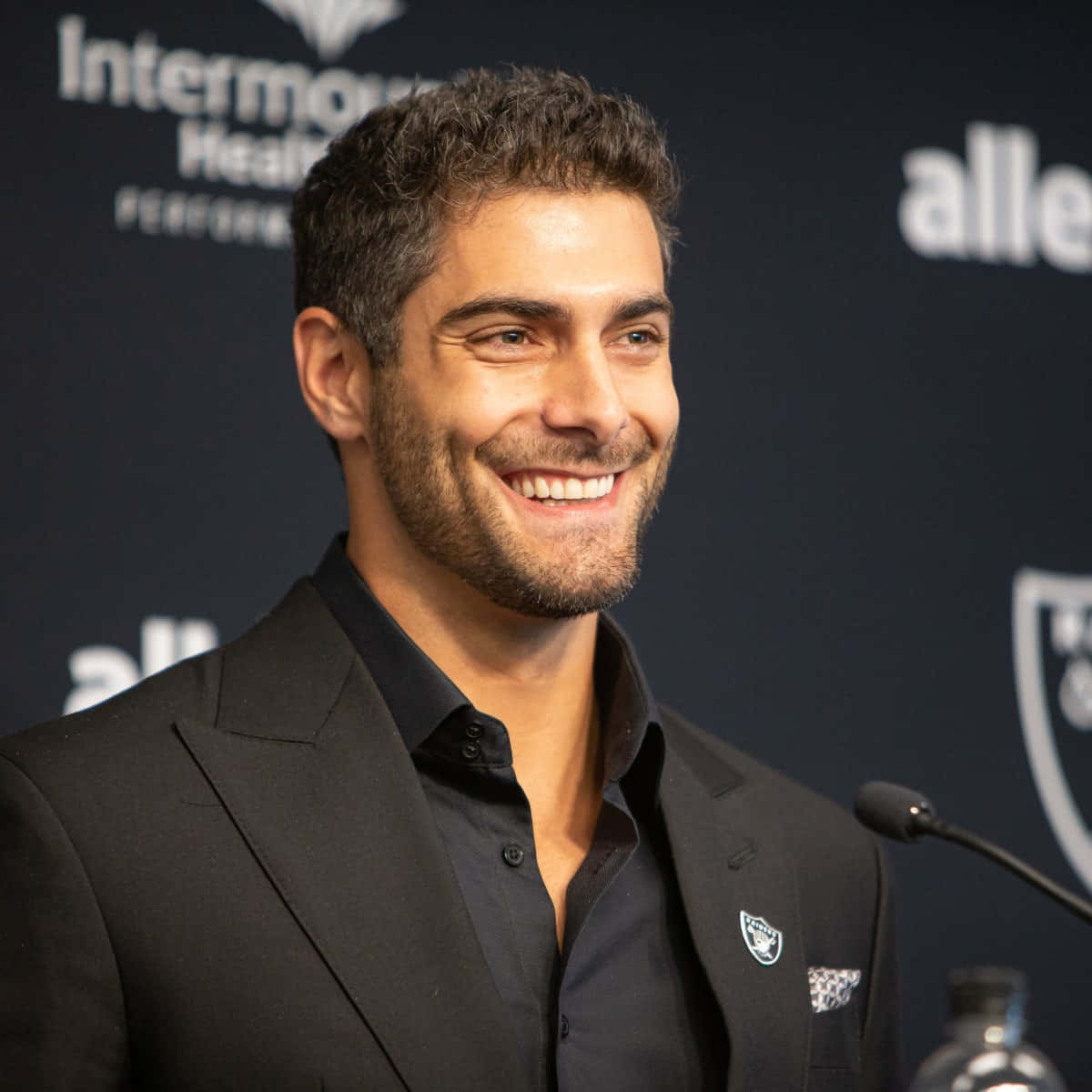Jimmy Garoppolo 桌布 壁紙 — 一位微笑的黑西裝和襯衫男士站在麥克風前，背景是深色品牌背景。