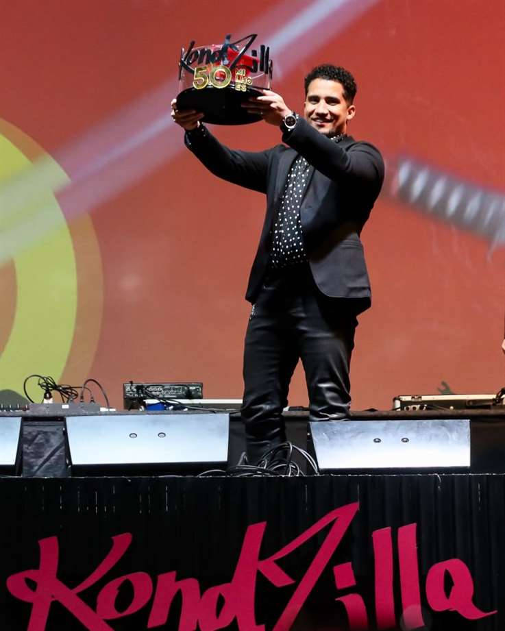 Trophy 桌布 壁紙 — 一位微笑的黑西裝男子在紅黃背景的舞台上高舉「KondZilla 50」獎盃。