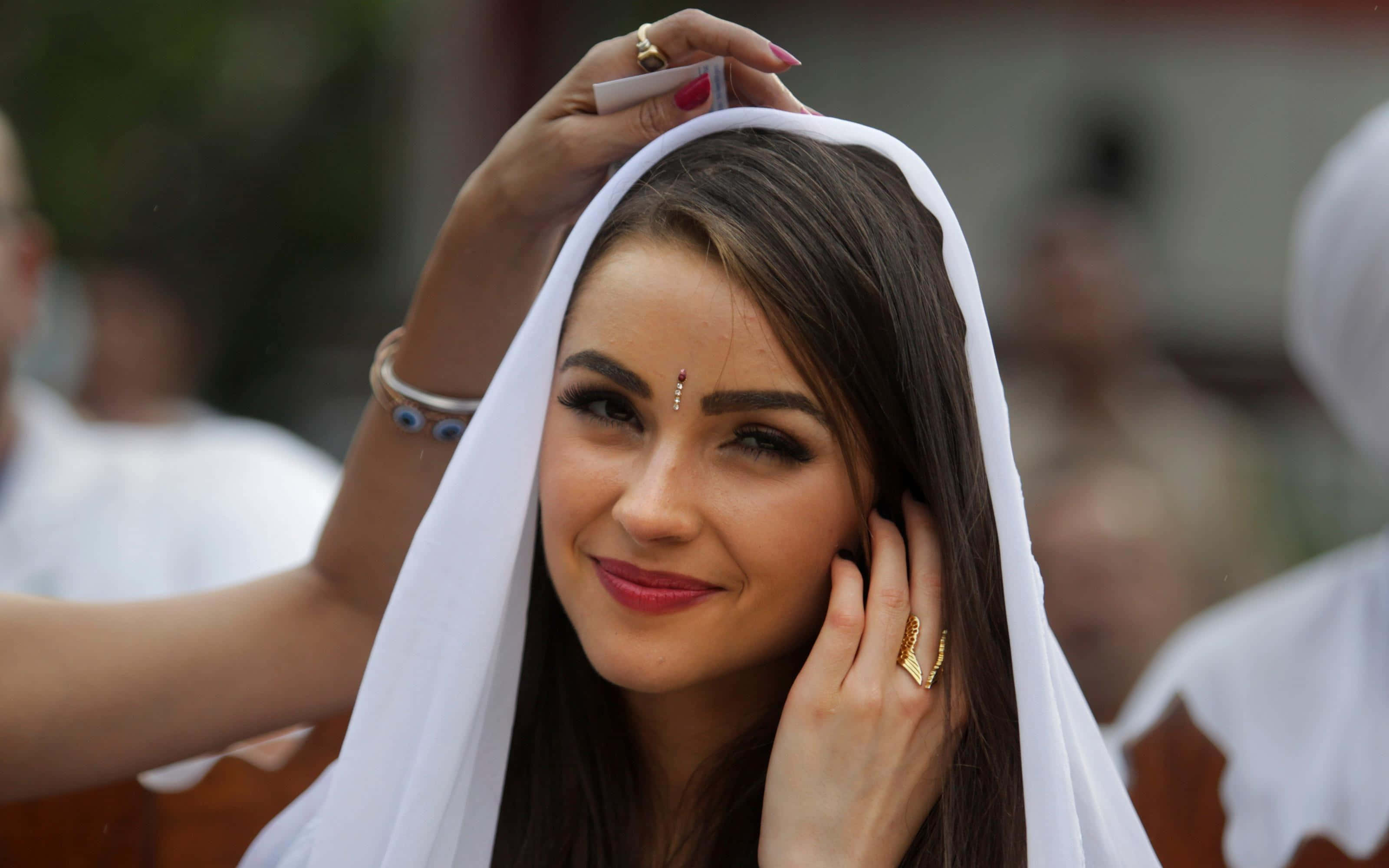 olivia culpo 桌布 壁紙 — 一位微笑的黑髮女子戴著白色頭紗，戴著金戒指和紅色唇膏，調整她的頭巾。