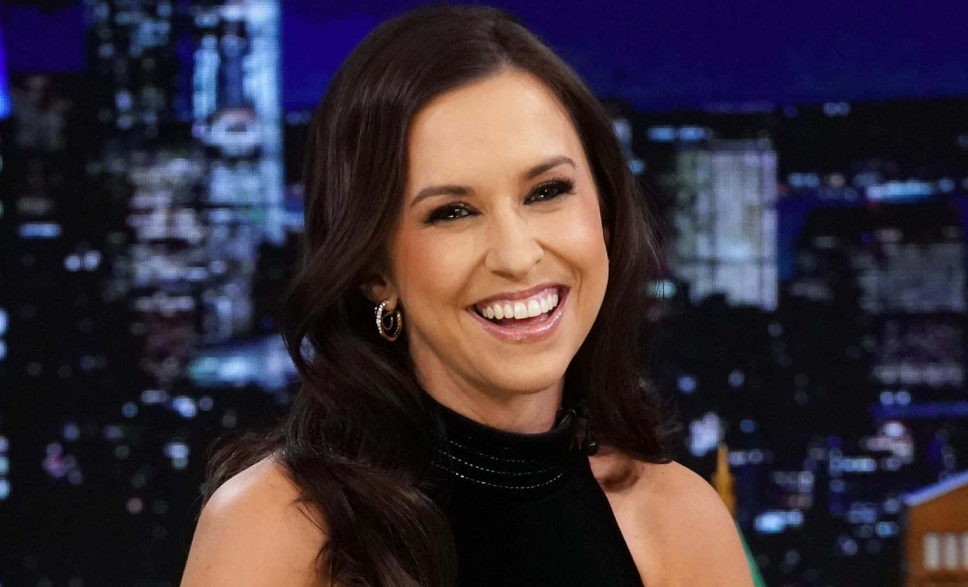 lacey chabert 桌布 壁紙 — 一位微笑的黑髮女性戴著圈圈耳環，穿著黑色上衣，背景是夜晚模糊的城市天際線。
